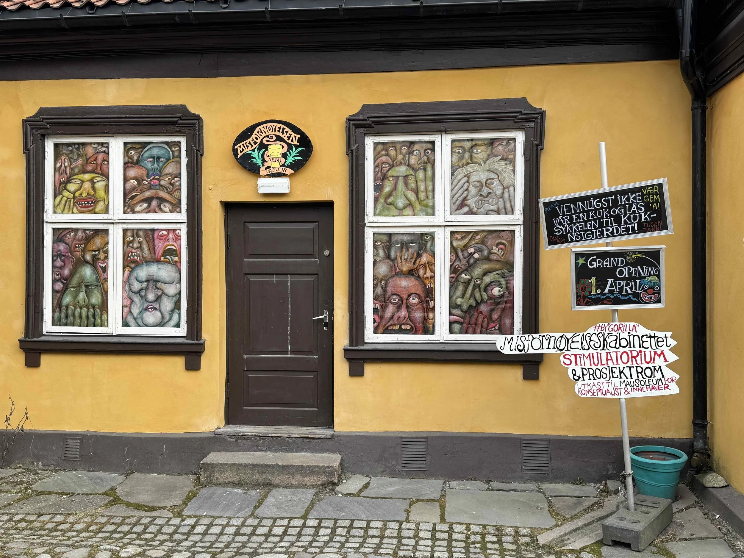 Exterior view of a yellow building with two stained glass windows featuring colorful, grotesque faces and figures. A dark brown door is between the windows, and there are signs advertising an opening event on April 1st and information in Norwegian ab