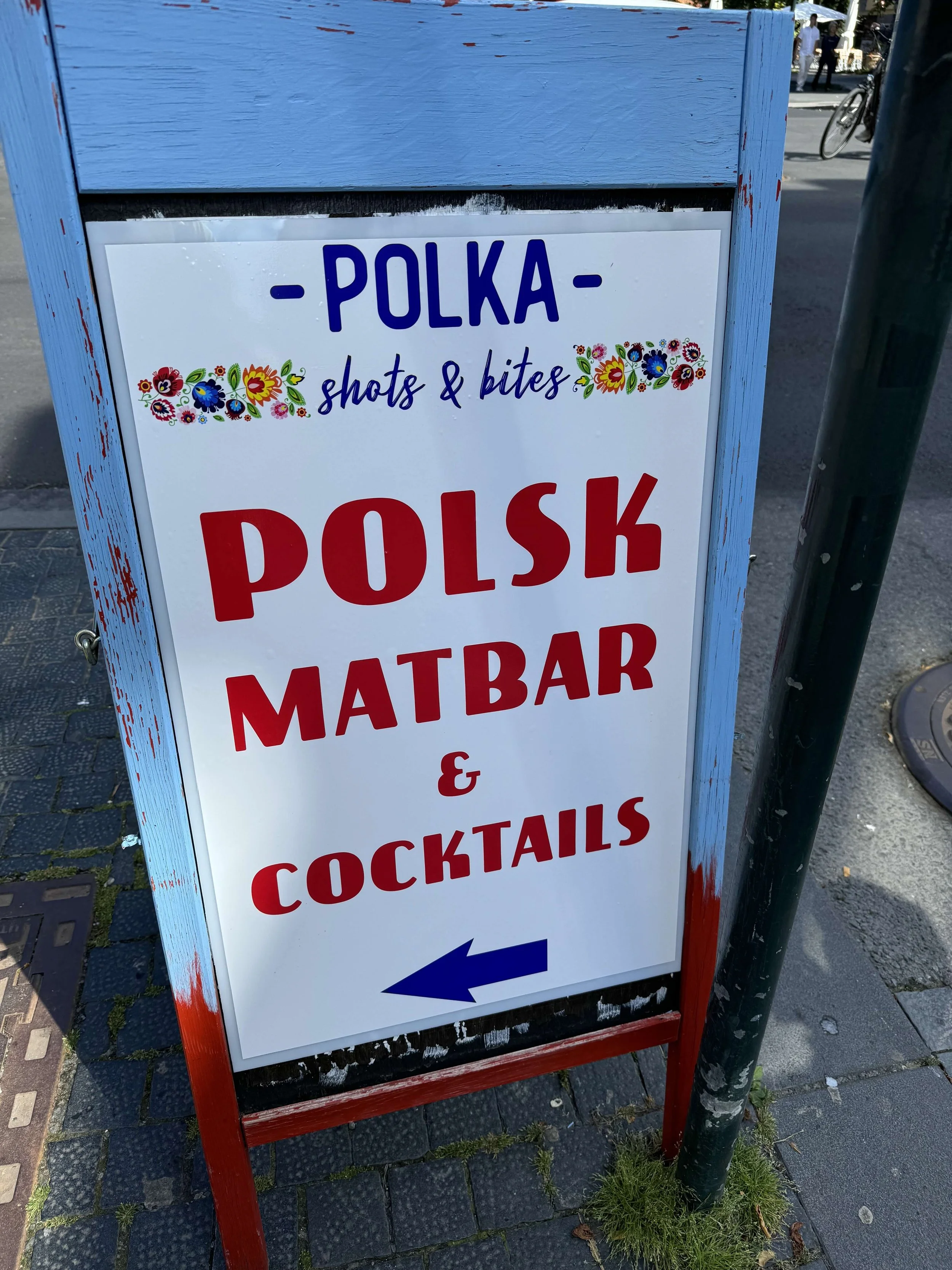 A sidewalk sign advertising a Polish food bar and cocktails, with an arrow pointing to the left.