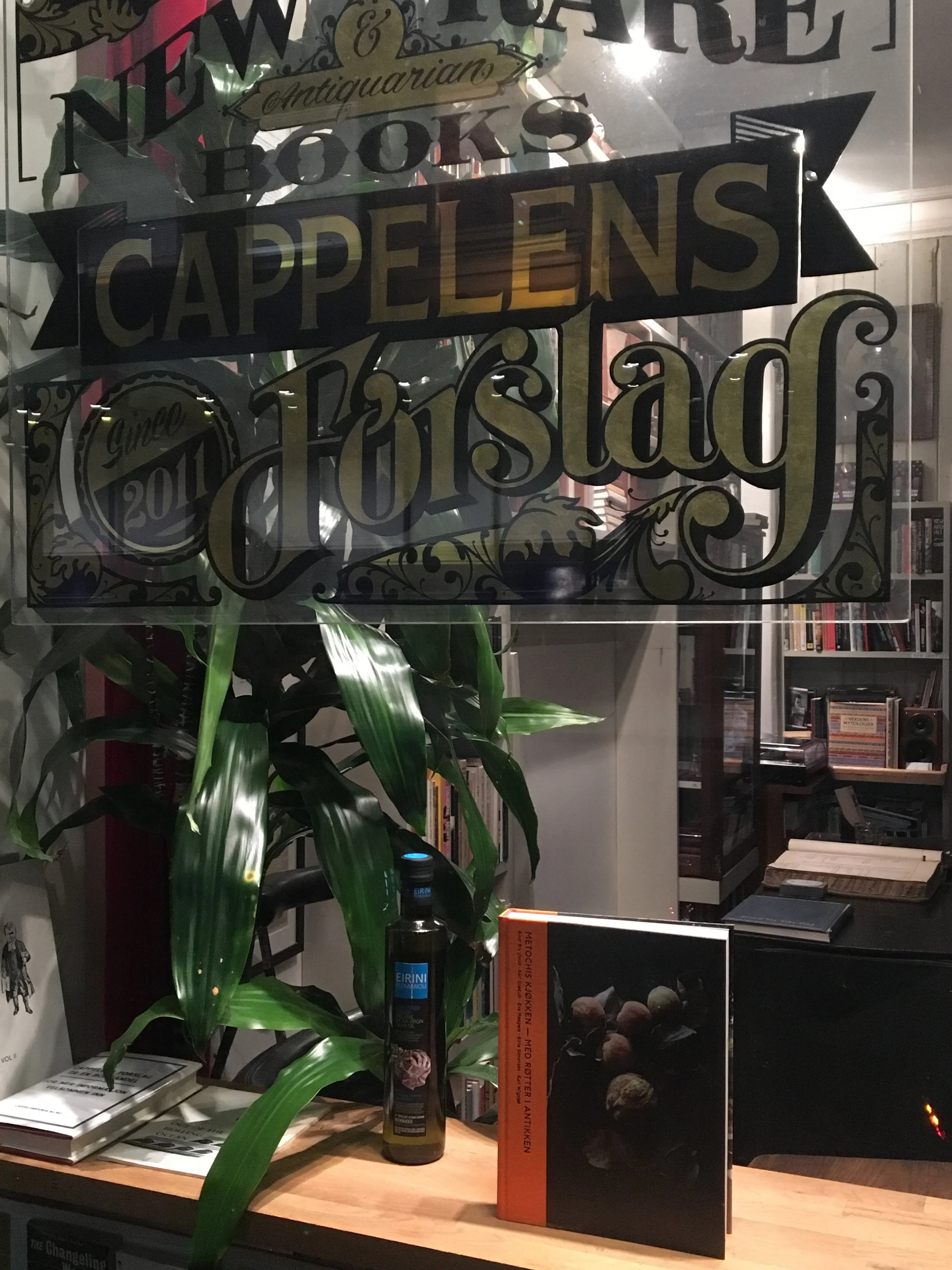 A glass sign on a window reads 'New & Rare Antiquarian Books Cappellens Torstagn.' Inside, there is a wooden table with books, a bottle, and a plant. Shelves with books are visible in the background.