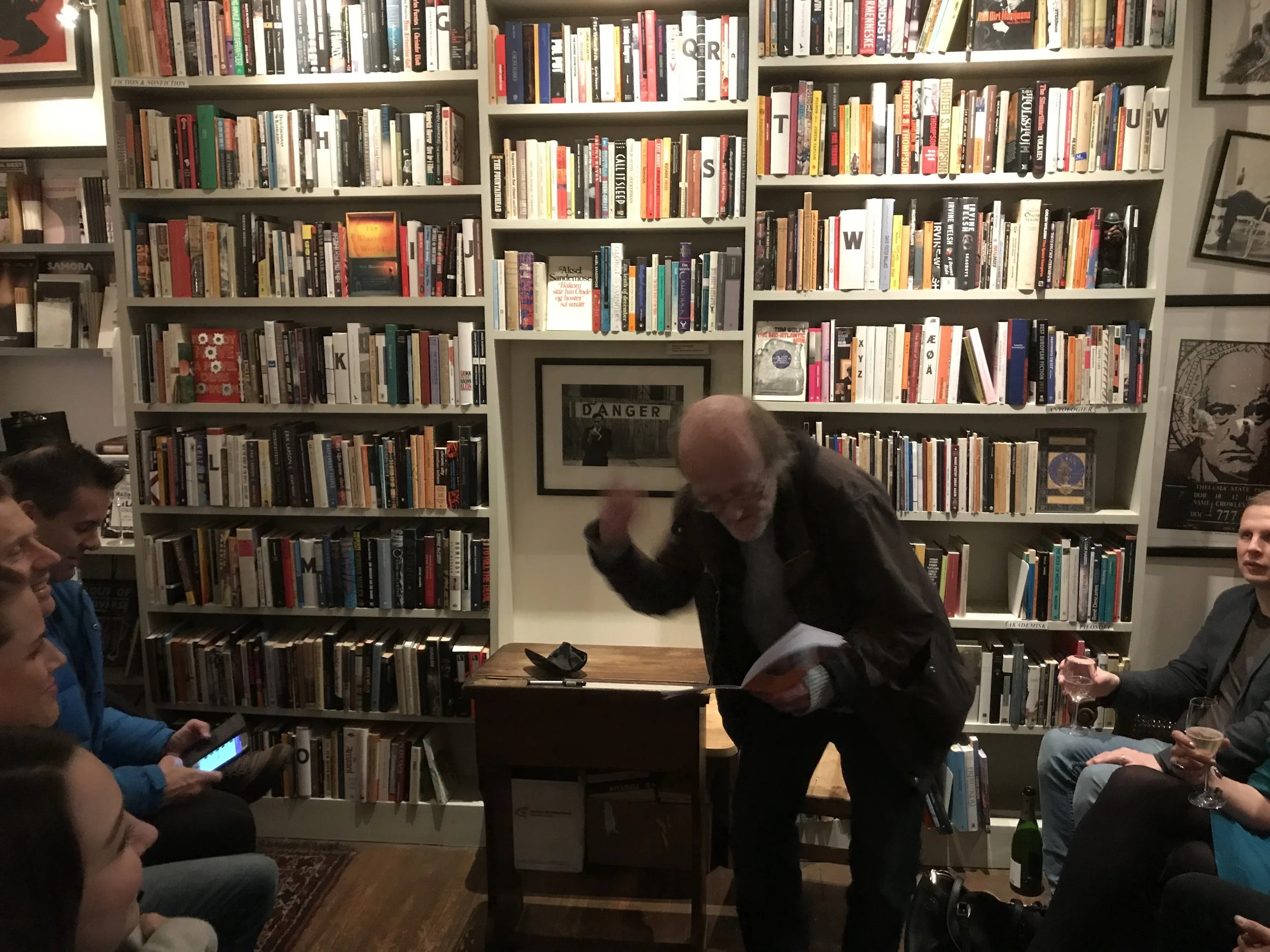 A man with glasses and gray hair standing and gesturing while reading from a book during a small presentation or reading event in a bookstore or library with tall bookshelves full of books. Several people are seated around him, some holding drinks, i