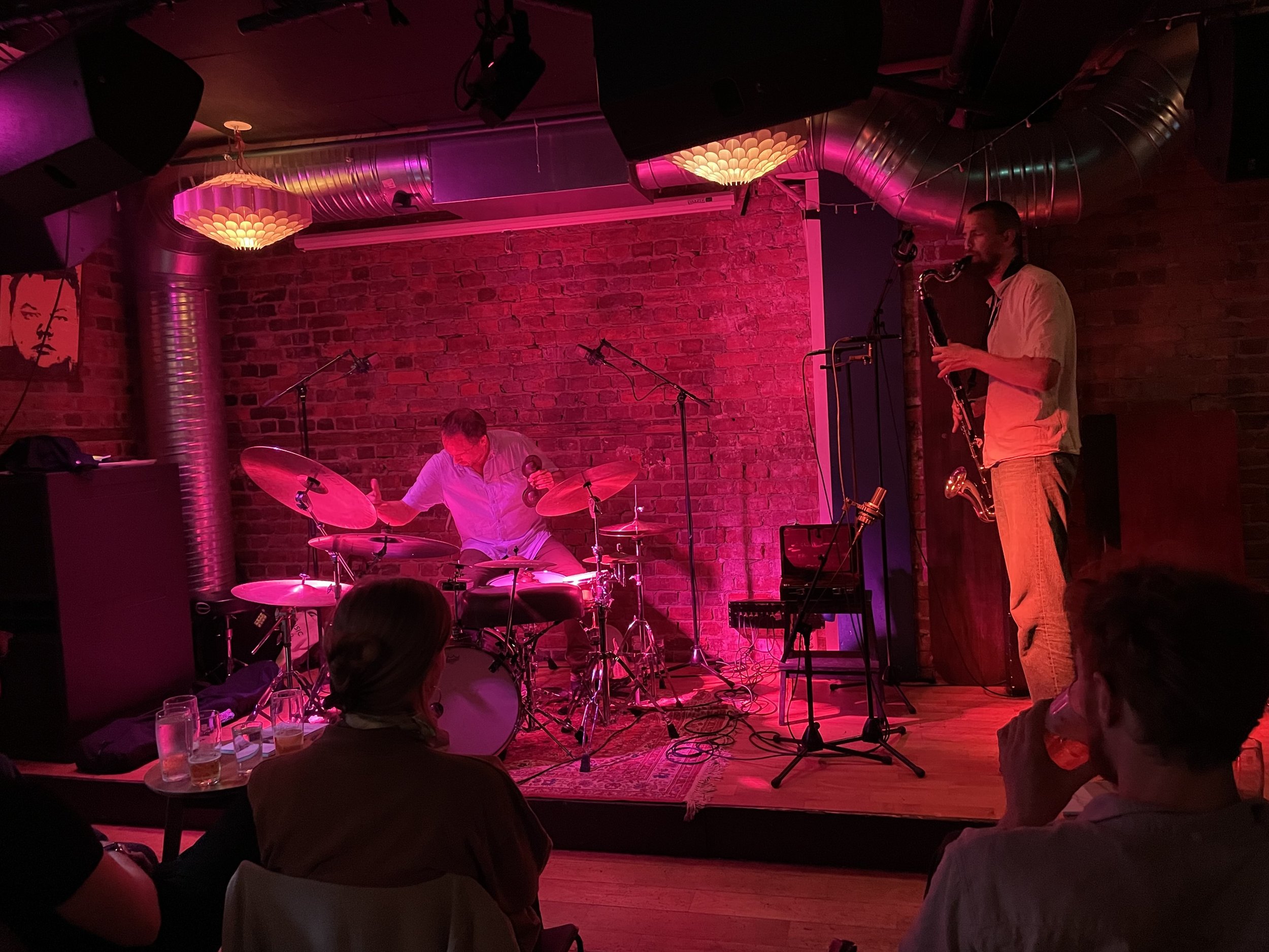Jazzy live music performance in a intimate indoor venue with brick wall background, featuring a drummer and a saxophonist, with audience members seated and enjoying the show.