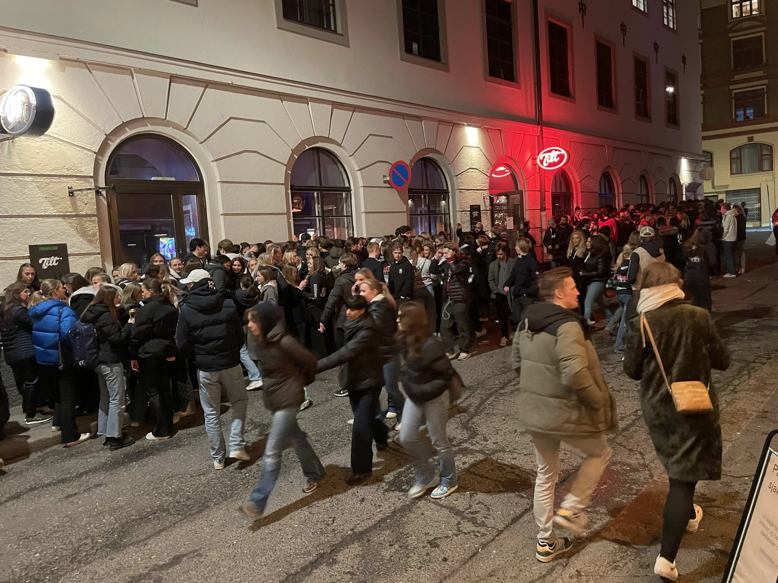 Night scene outside a crowded nightclub with long lines of people waiting to enter. The building has arched windows, white walls, and illuminated signs, including a red neon sign.