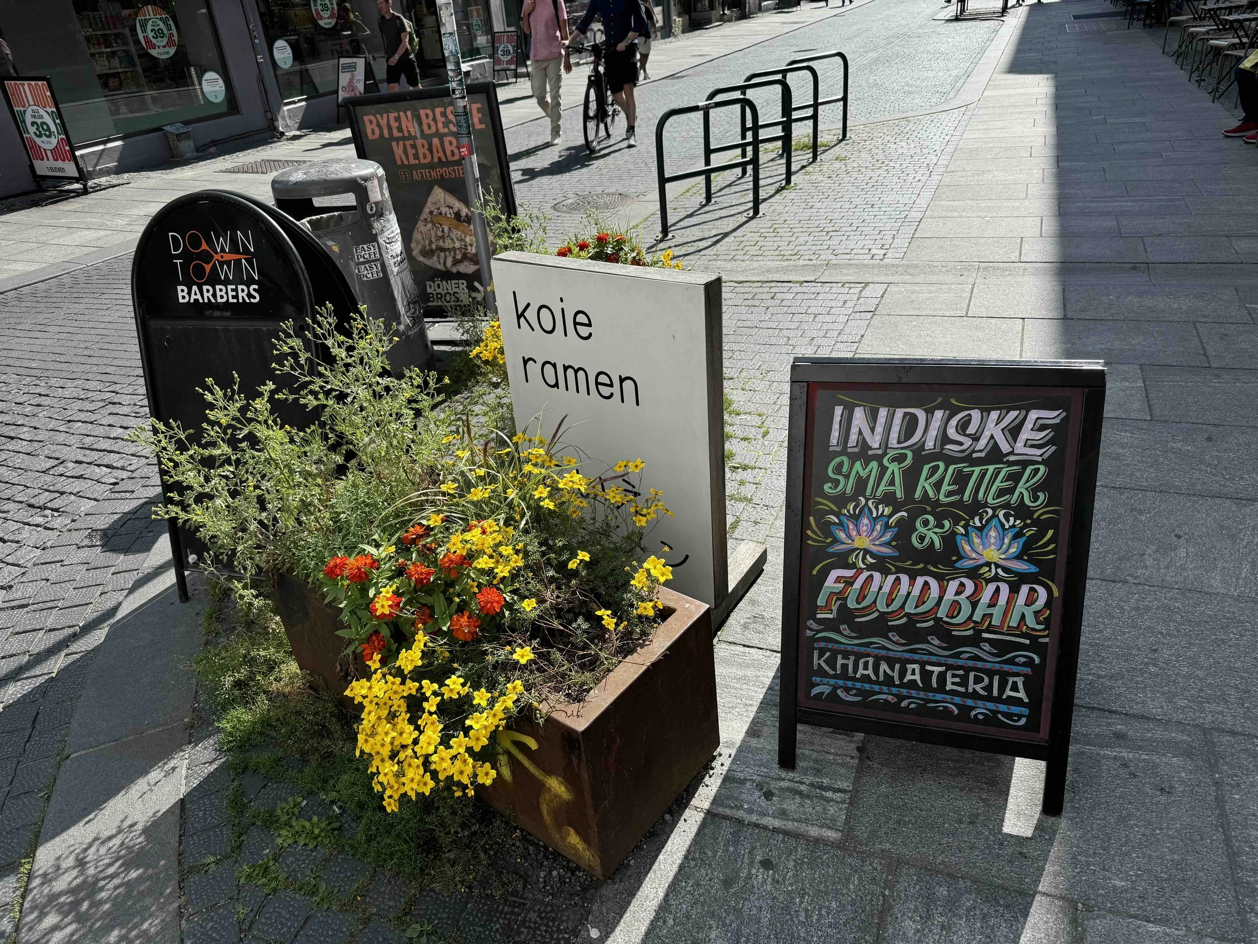 Street sidewalk with colorful flowers in a planter, chalkboard and signboards advertising a food bar, ramen, and a barber shop, with pedestrians walking in the background.