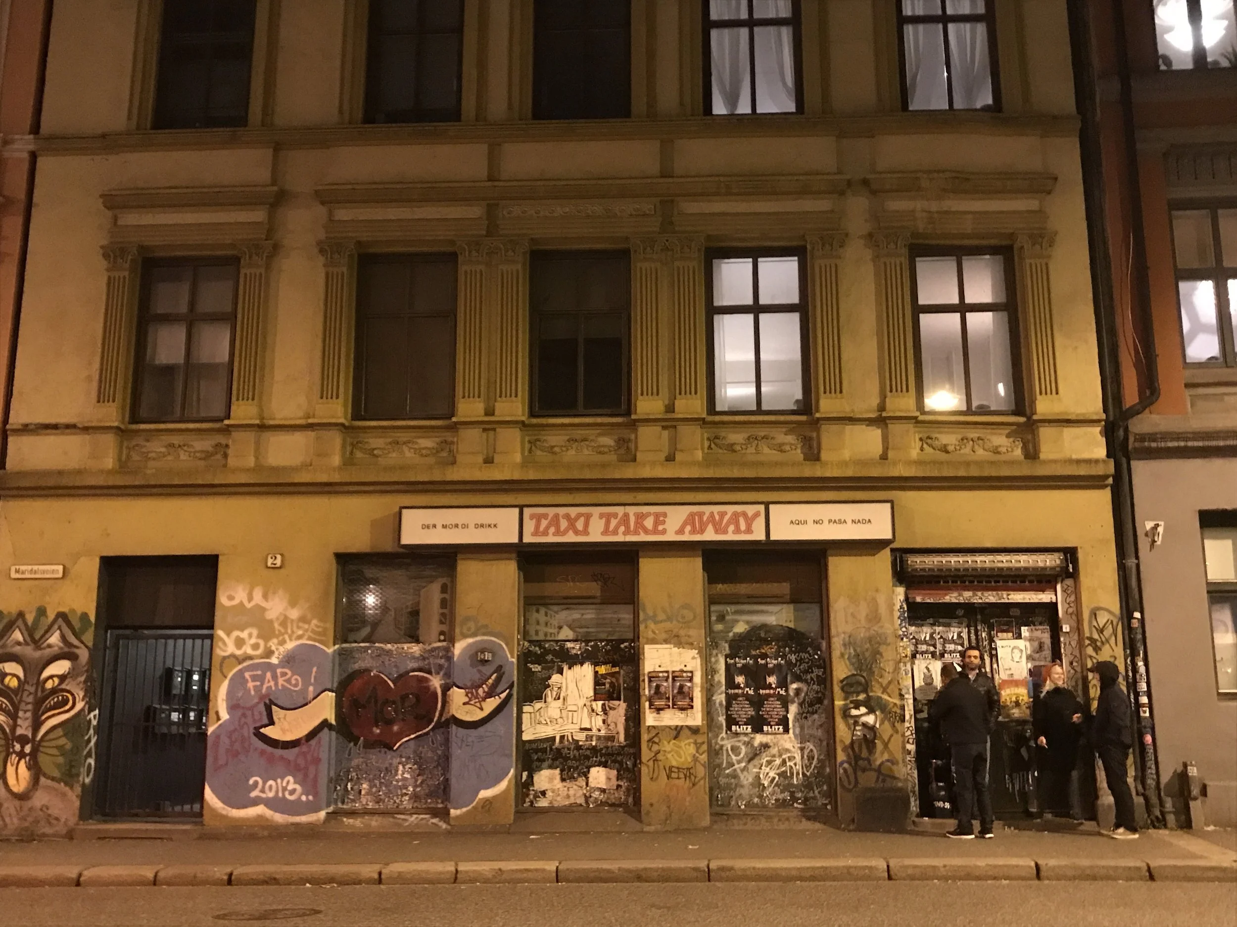 Nighttime street scene with a yellow building featuring graffiti, a sign reading 'Taxi Take Away,' and a group of four people standing outside.