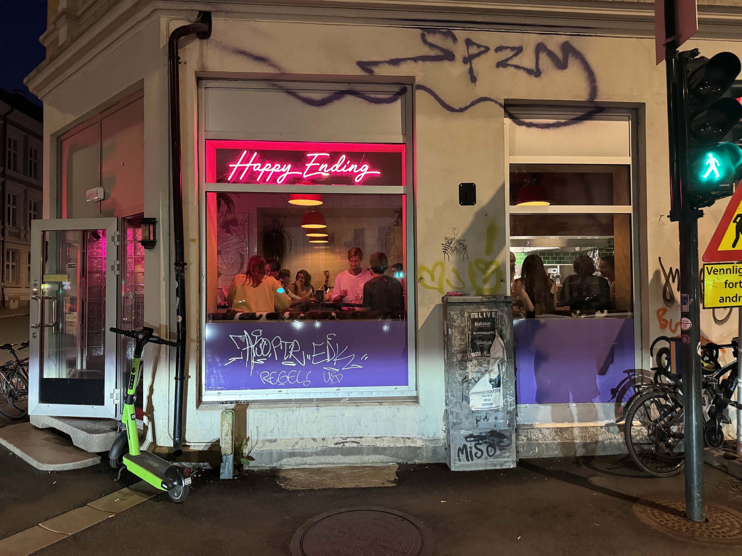 Night view of a street corner with a cafe that has a neon sign reading 'Happy Ending'. Inside, people are sitting at tables, with some visible through the large windows. The building's exterior has graffiti, and there are bicycles and an electric sco