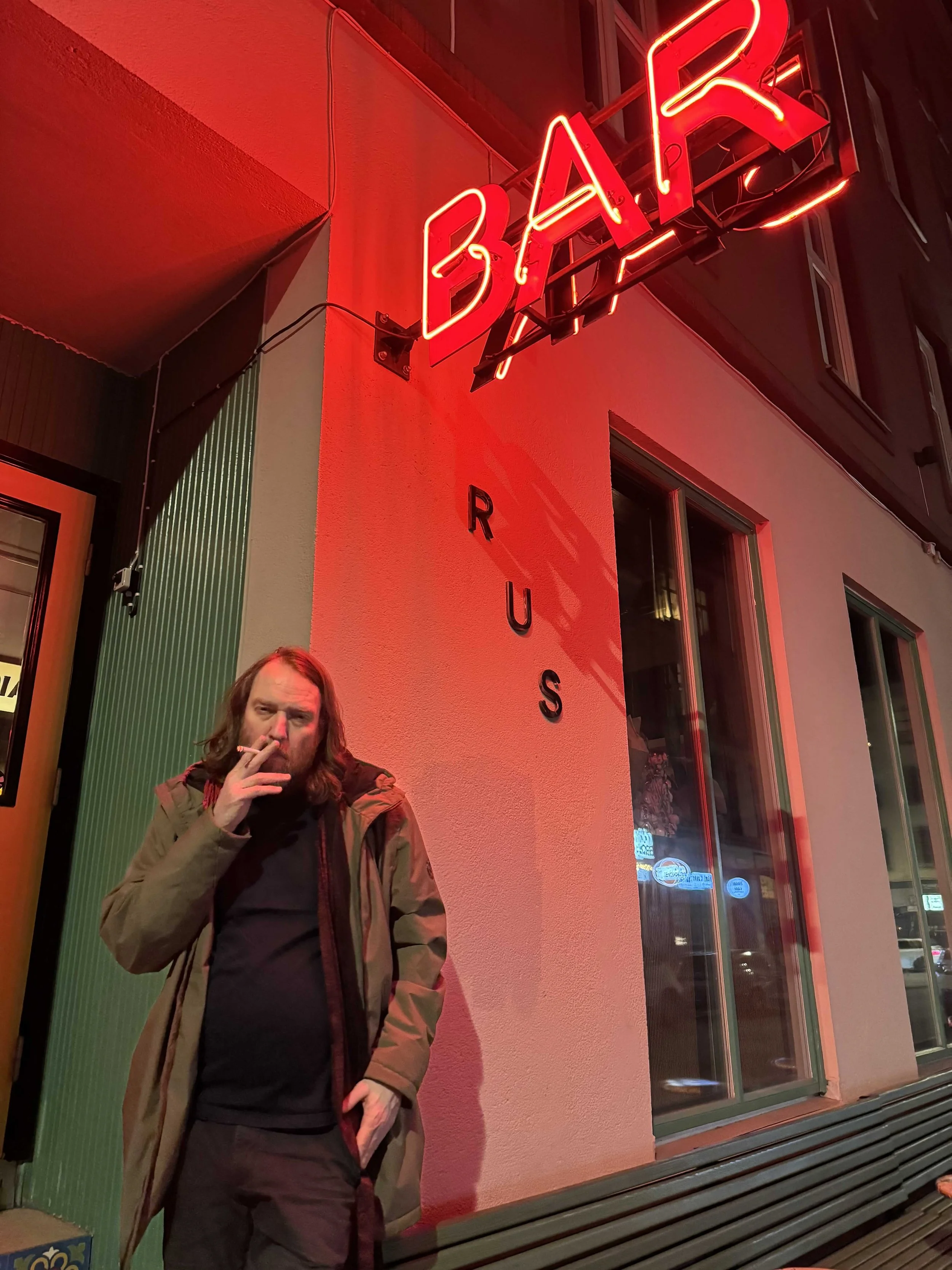 A man with long hair, wearing a brown jacket and black shirt, stands outside a burger restaurant at night, smoking a cigarette. The restaurant has a bright red neon sign that reads "BURGER" and the word "RUS" vertically on the wall.