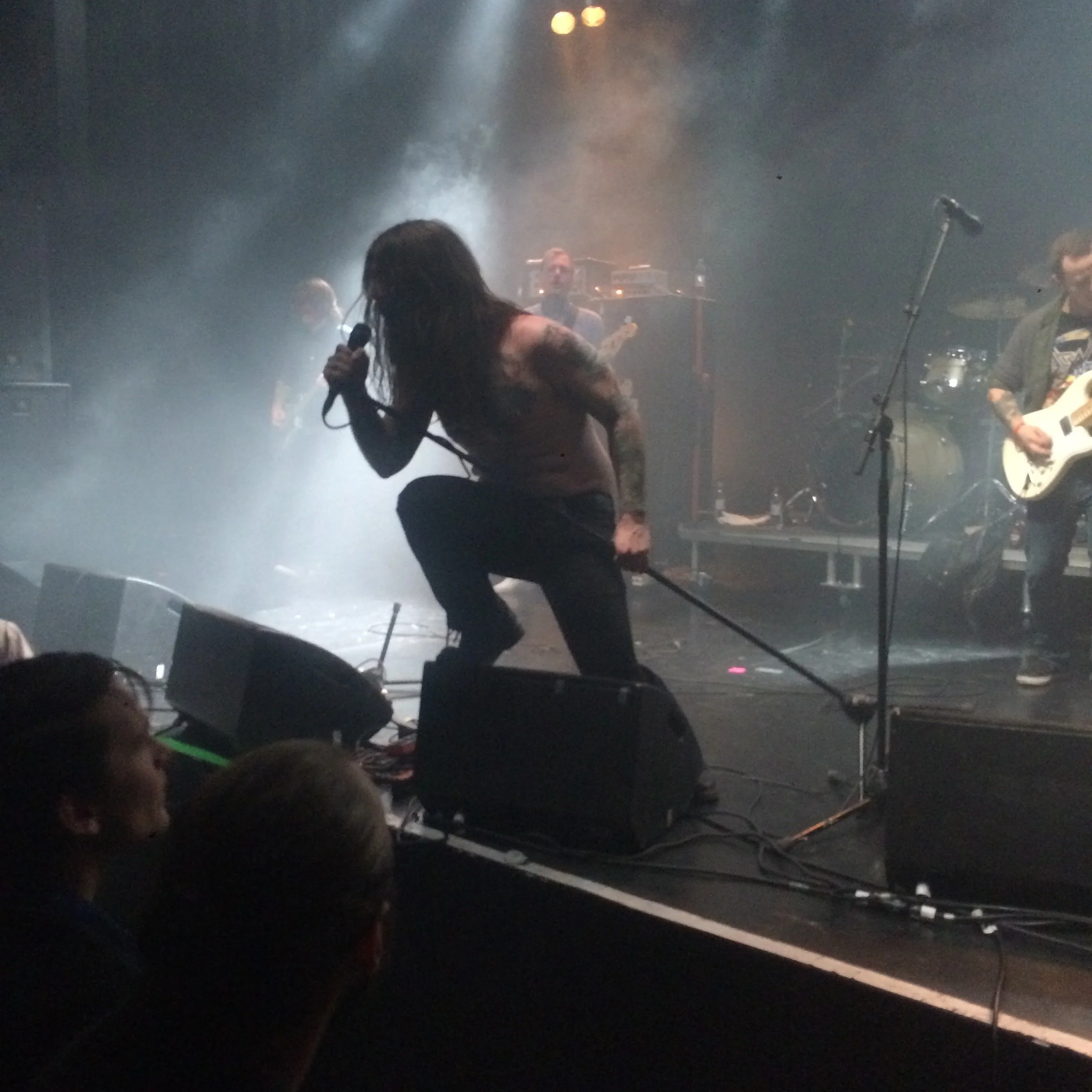 A band performing on a smoky stage with a singer crouching and holding a microphone, surrounded by members playing guitars and drums.