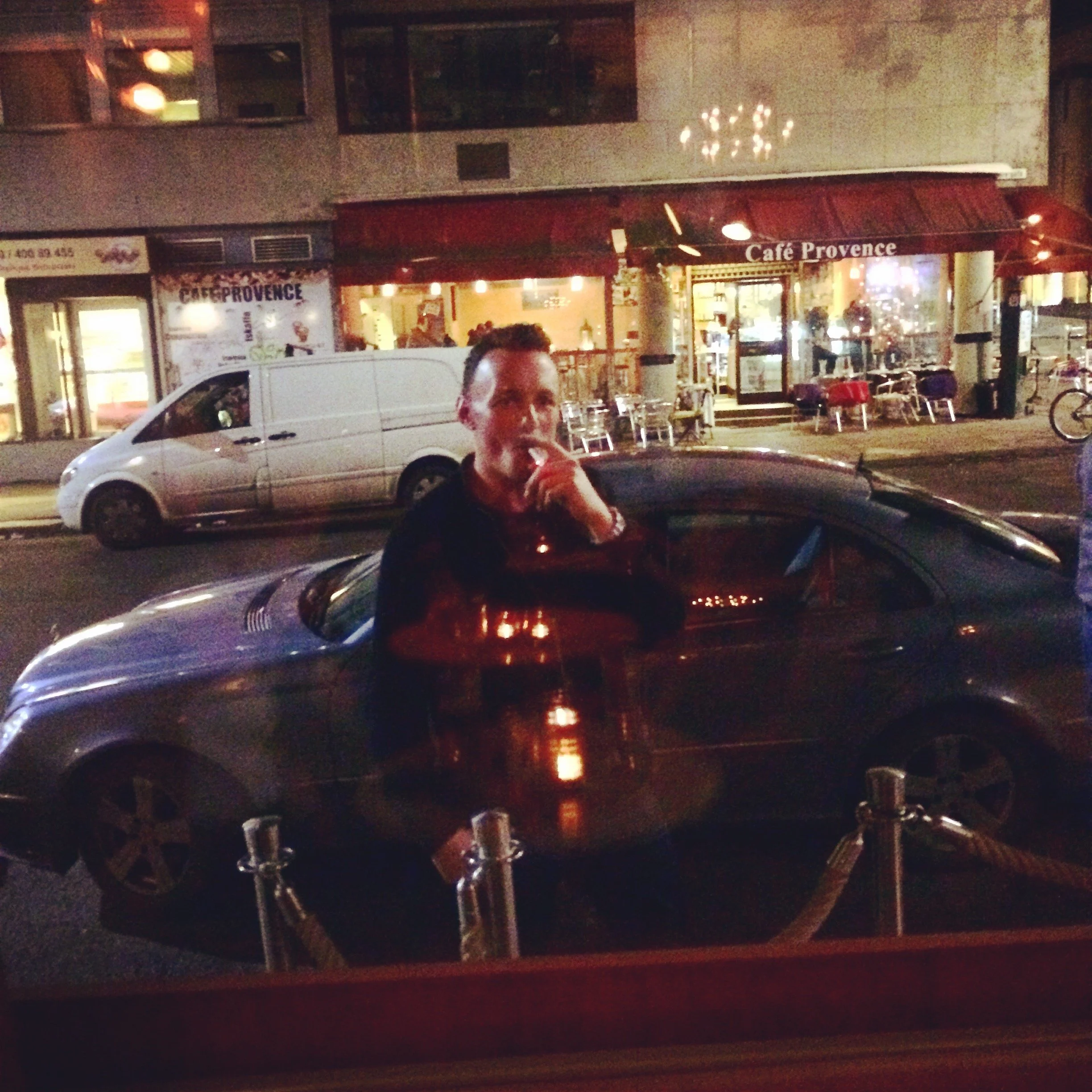 A man standing outside a restaurant at night, smoking a cigarette. There is a silver car in front of him and a white van on the street behind. The restaurant has a red awning with the sign 'Cafe Provence' and outdoor seating.