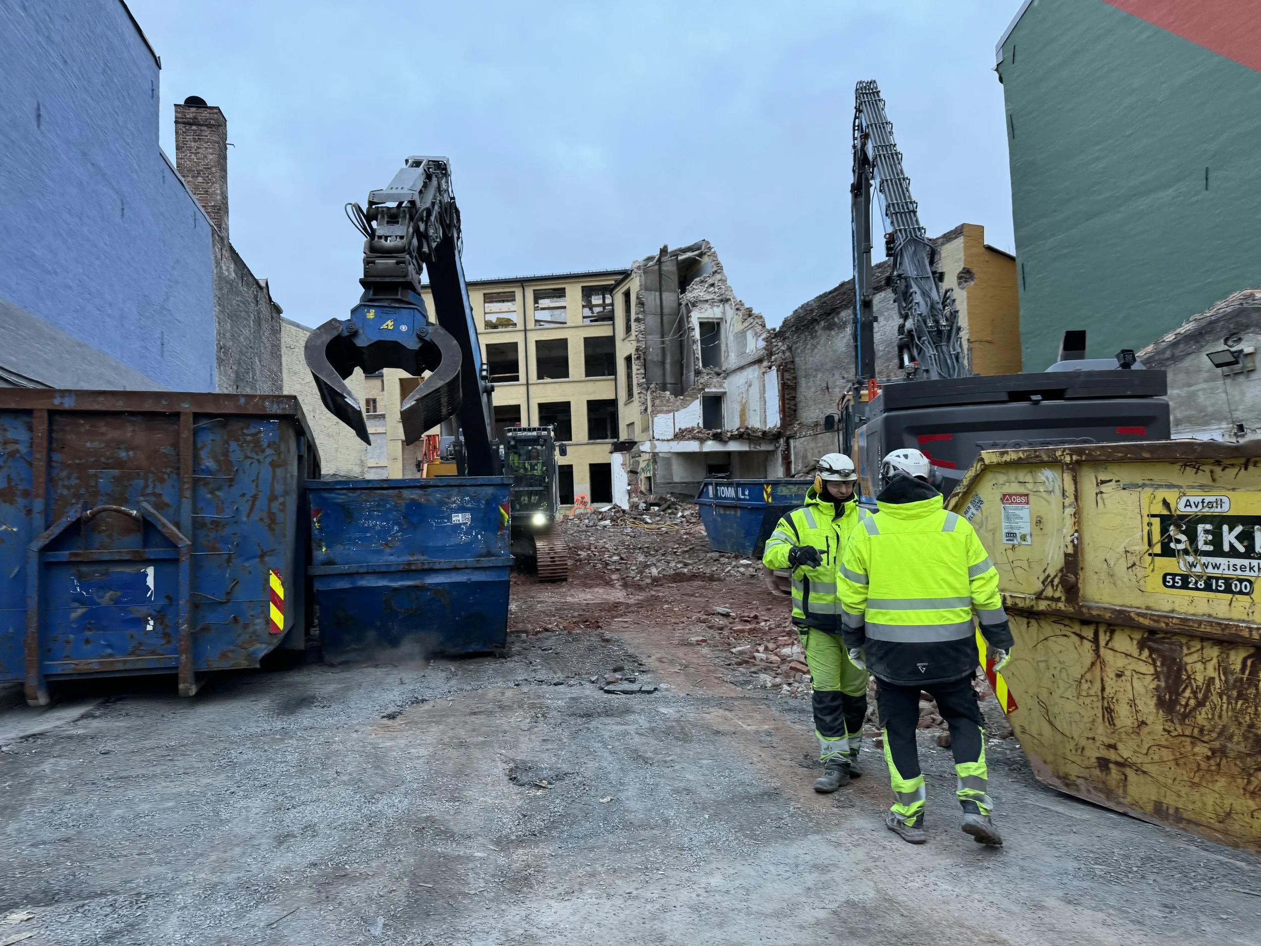 Construction workers in neon safety jackets and helmets standing on a dirt road, operating heavy machinery to demolish a building in an urban area.