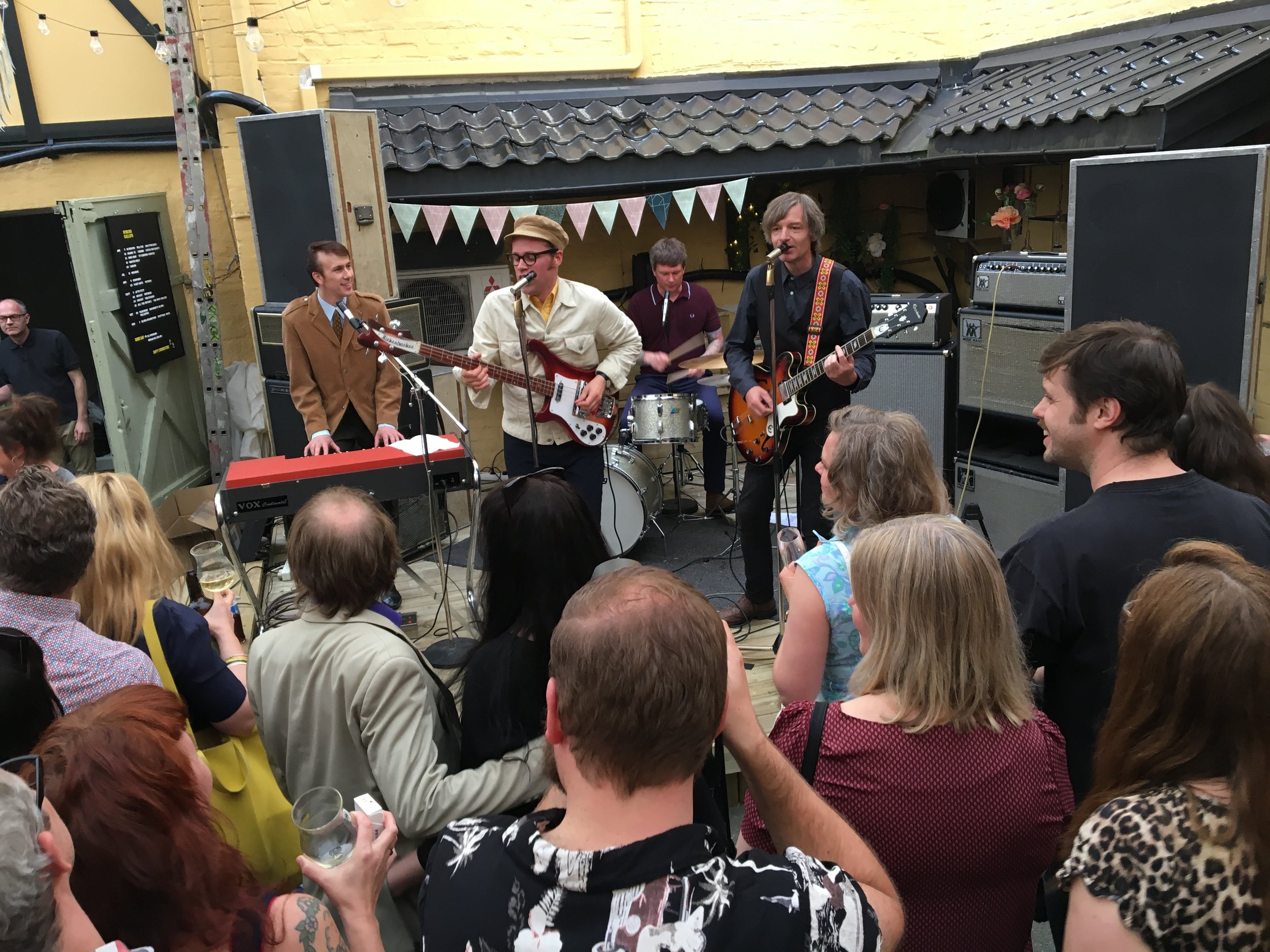A band performing live outdoors with an audience watching, including a keyboardist, guitarist, bassist, and drummer, in a cozy setting with string lights and bunting.