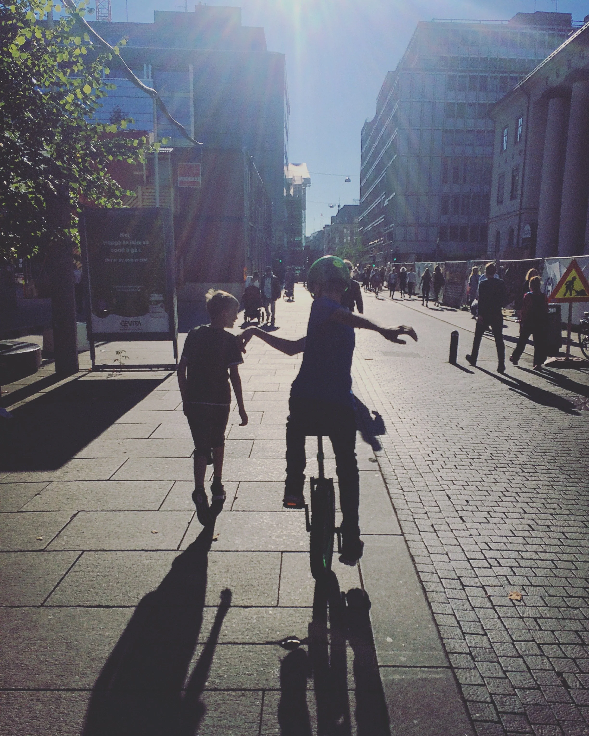 People walking on a city sidewalk in bright sunlight, with a child riding a bicycle and holding hands with another child, surrounded by tall buildings and street signs.
