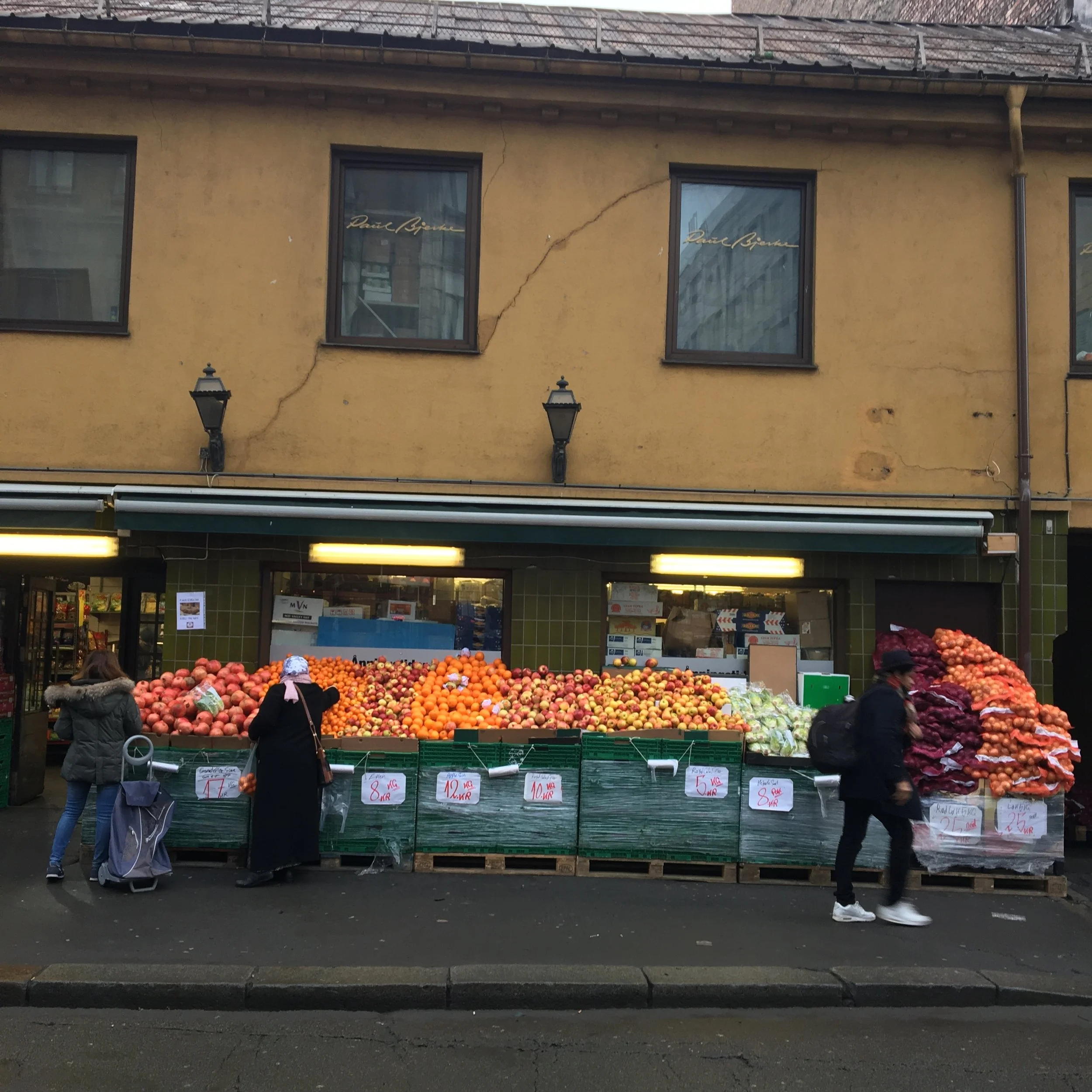 Street market with fresh produce, including apples and onions, in front of a yellow building with windows and two outdoor lamps.