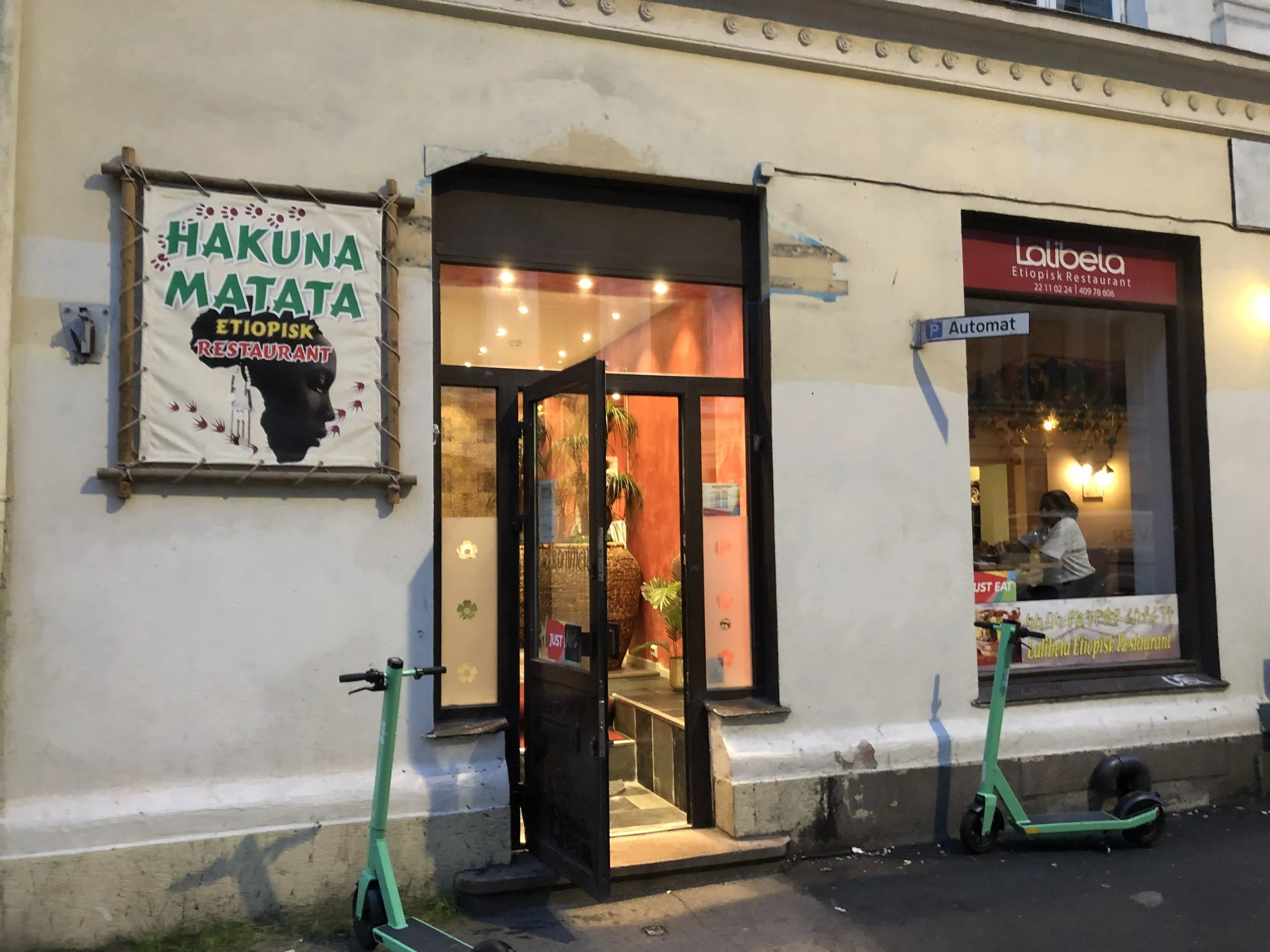 Exterior view of a restaurant named Hakuna Matata, featuring a colorful sign with a stylized face and the text 'Hakuna Matata Etiopisk Restaurant.' The entrance has a glass door, and there are two electric scooters parked outside. Inside, a person is