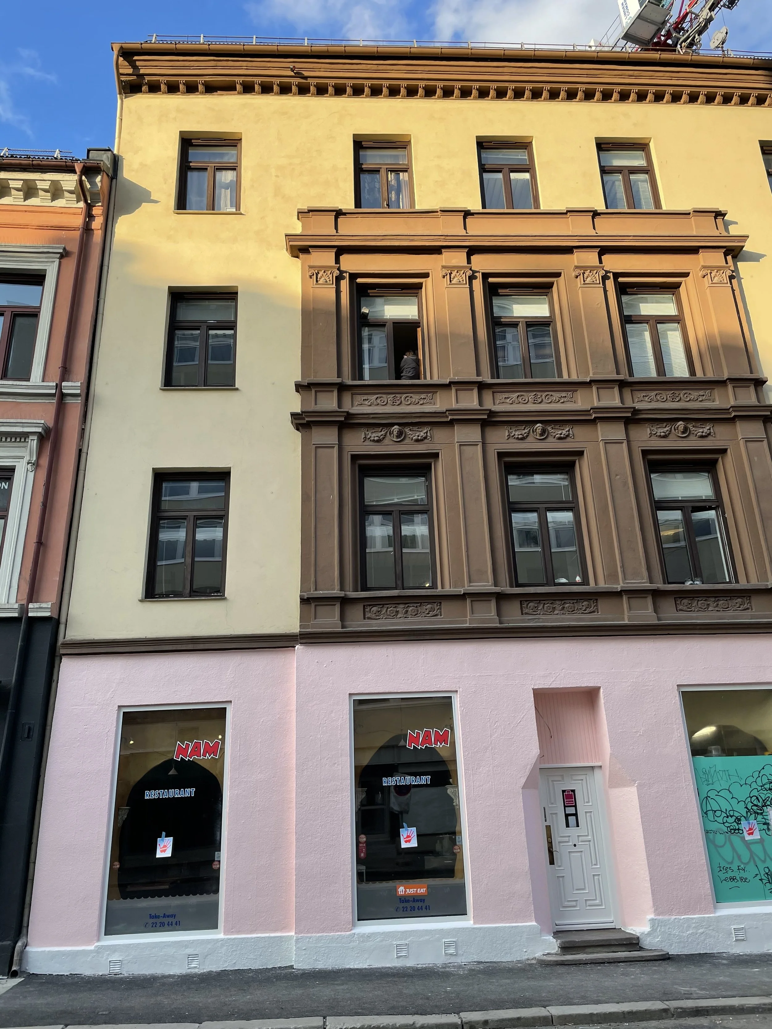 A multi-story building with storefronts on the ground level and residential or office spaces above. The building has a pink lower facade with two large windows displaying signs for a restaurant called NAM, and a white door in the middle. The upper le