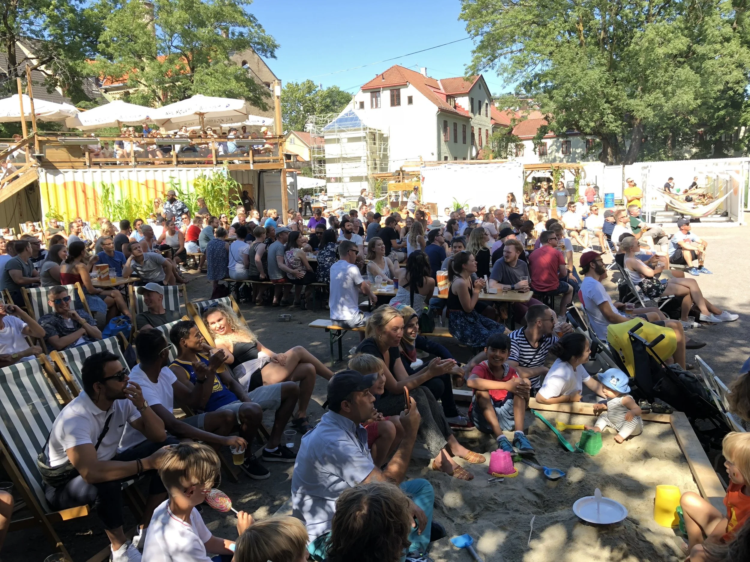 Crowd of people watching a performance or event outdoors, with some children playing in a sandbox in the foreground.