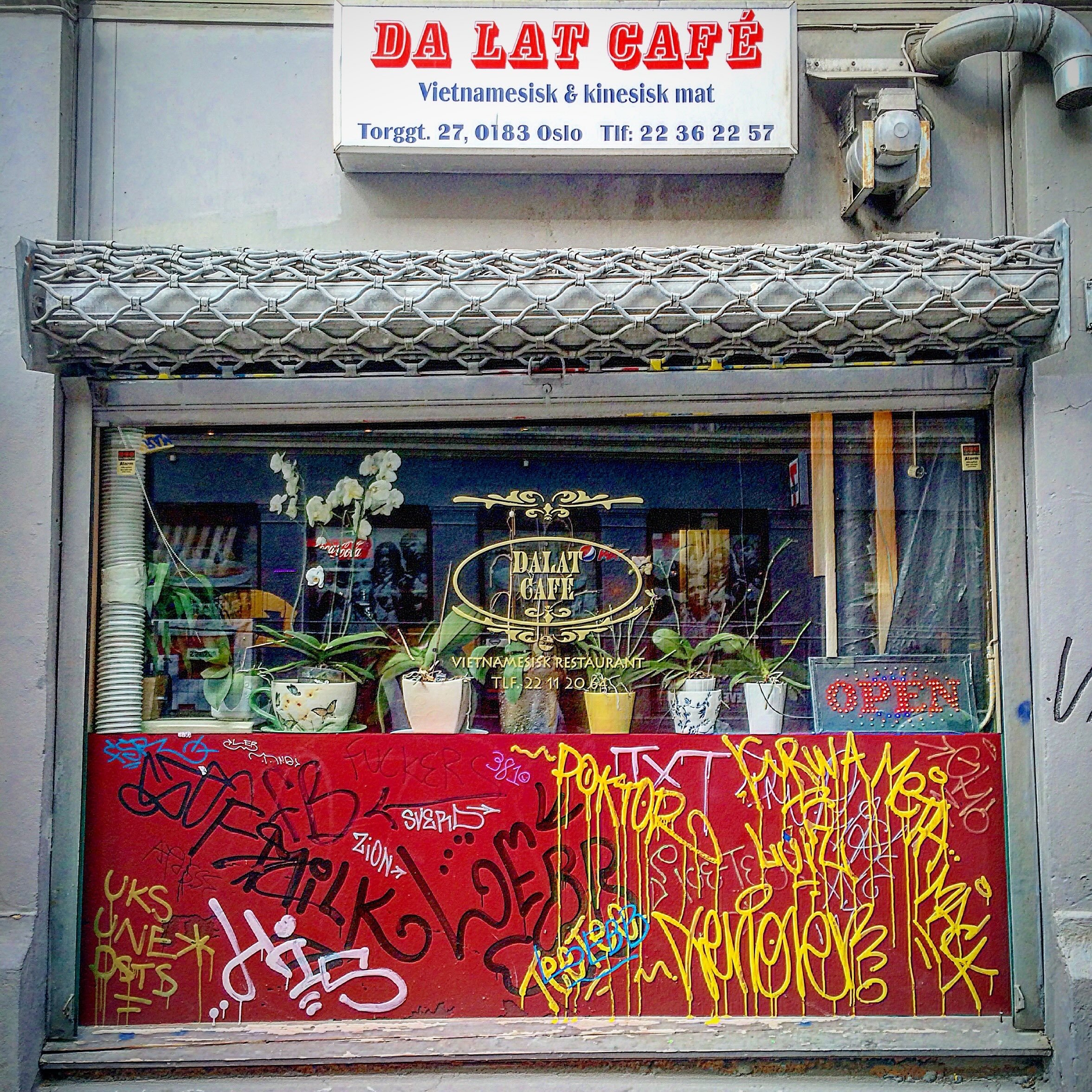 Street view of a Vietnamese and Chinese restaurant named Da Lat Café in Oslo. The storefront has graffiti on the red lower panel, a glass window display with potted plants, and a sign indicating the restaurant is open.