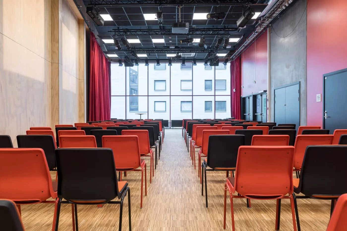 Empty conference room with red and black chairs facing a stage with large windows and red curtains, with a city view outside.
