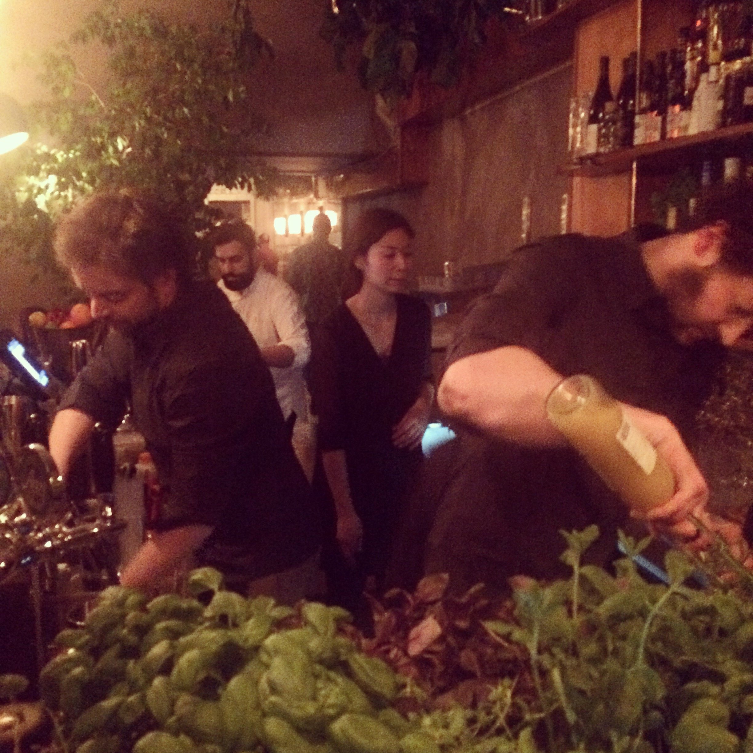People working behind a bar at a dimly lit restaurant or bar, with one person pouring a drink and others observing.