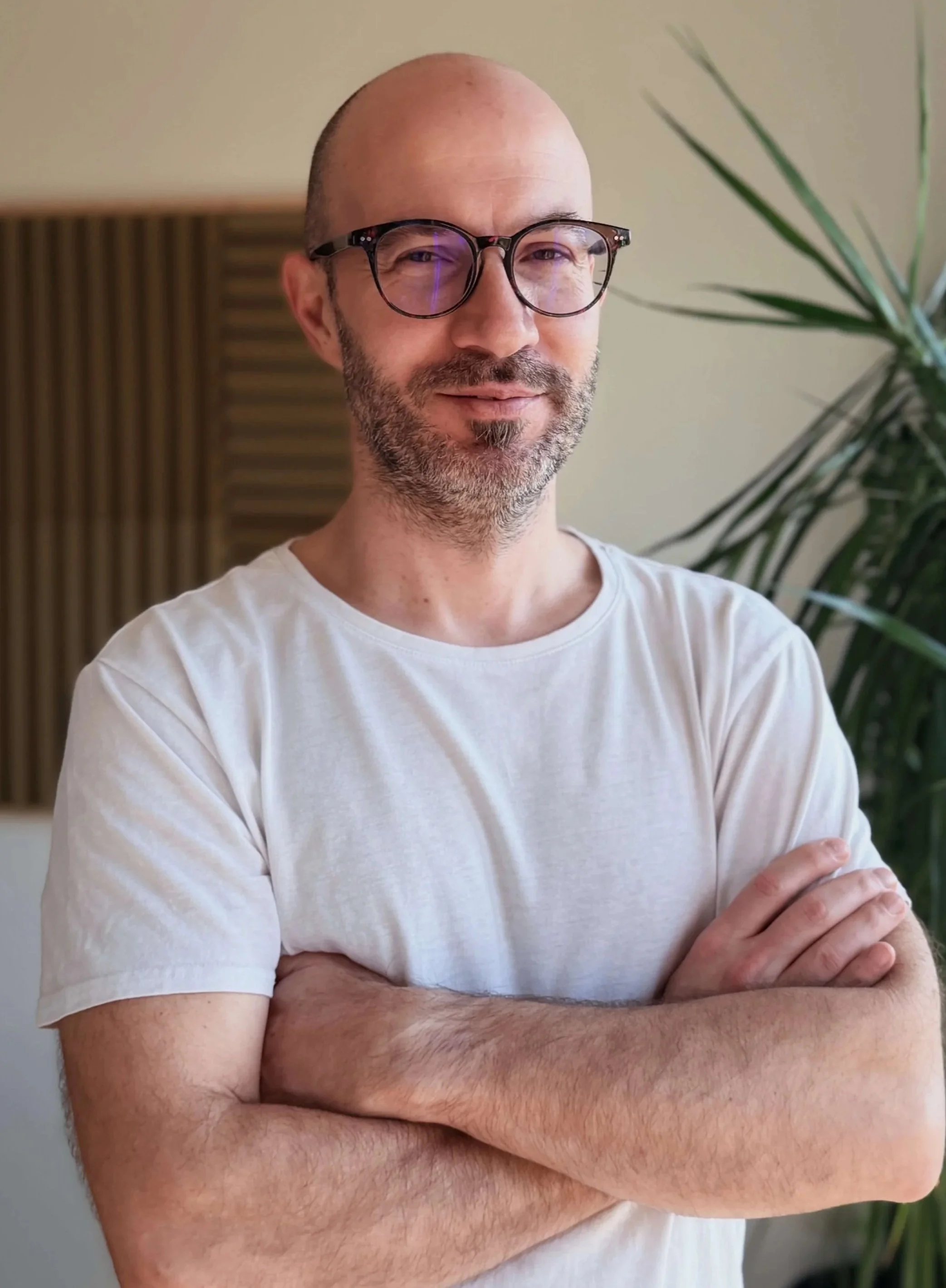 Muškarac s naočalama i bradom, u bijeloj majici, stoji prekriženih ruku u zatvorenom prostoru pored velike zelene biljke.