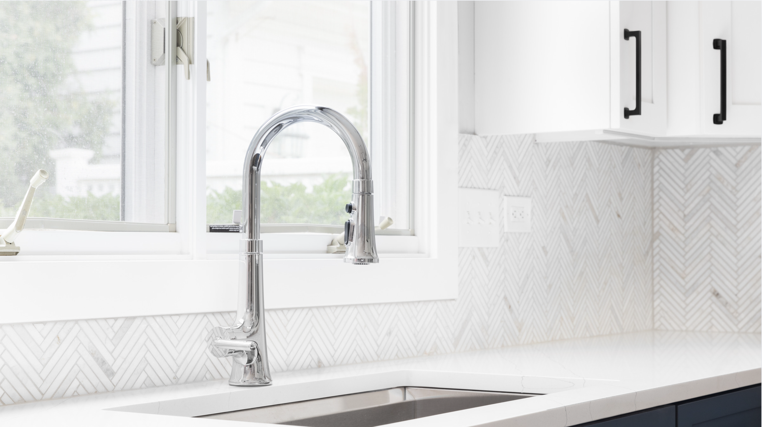 Kitchen sink with a chrome faucet in front of a window, white cabinetry, and a patterned backsplash.