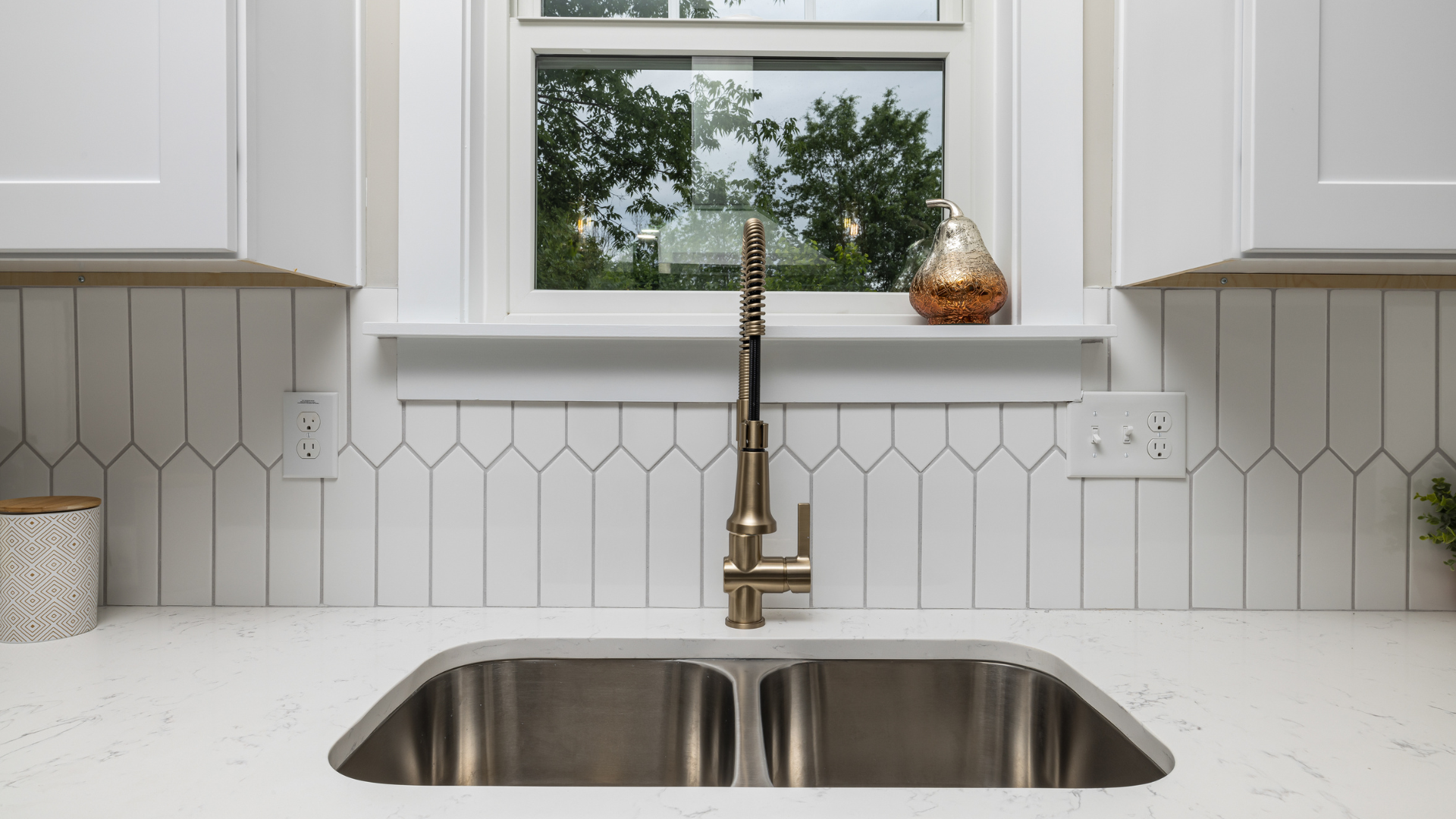 Kitchen sink with a modern gold faucet, white countertops, and white hexagonal tile backsplash. There is a window above the sink showing green outside trees, and a small decorative vase on the windowsill. Two electrical outlets are visible on either side of the sink.