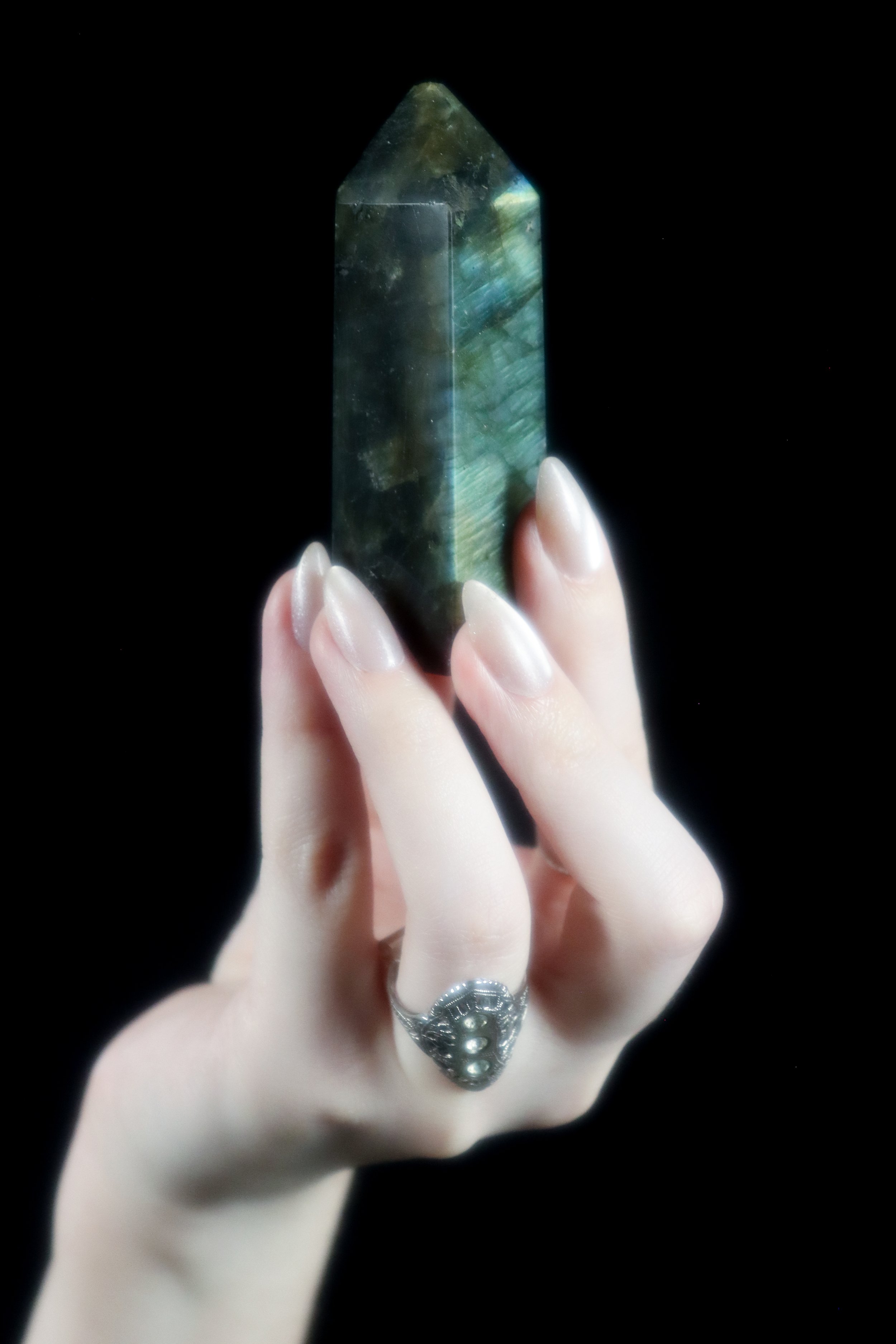 A hand with a silver ring holding a large, labradorite tower against a black background.
