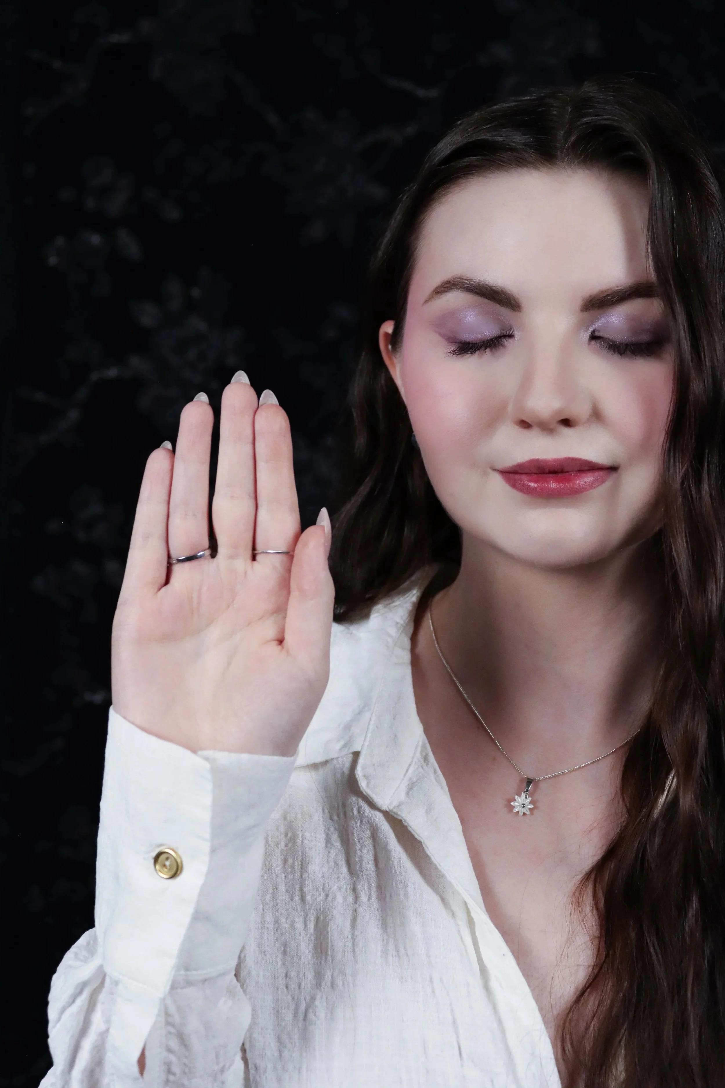 A woman with long dark hair, closed eyes, wearing a white shirt with gold button details, a dainty necklace with a flower-shaped pendant, and holding her hand up with fingers spread apart to give Reiki.