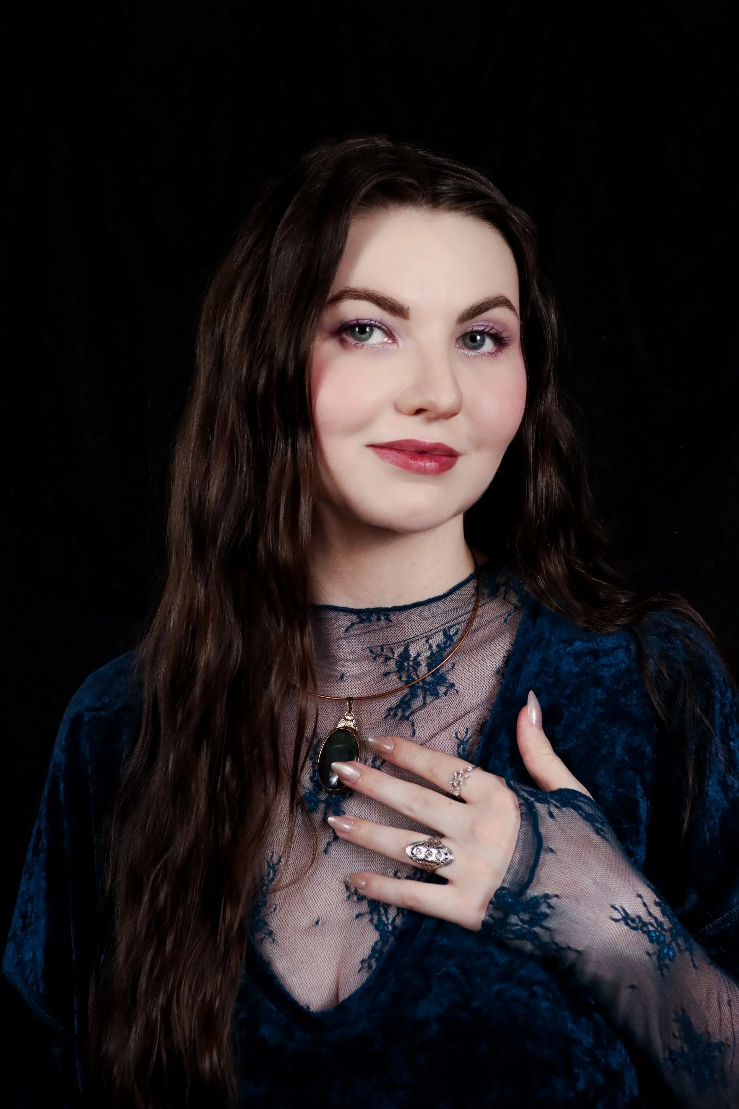 Portrait of a woman with long brown hair, wearing jewelry, a dark blue lace and velvet top, standing in front of a black background.