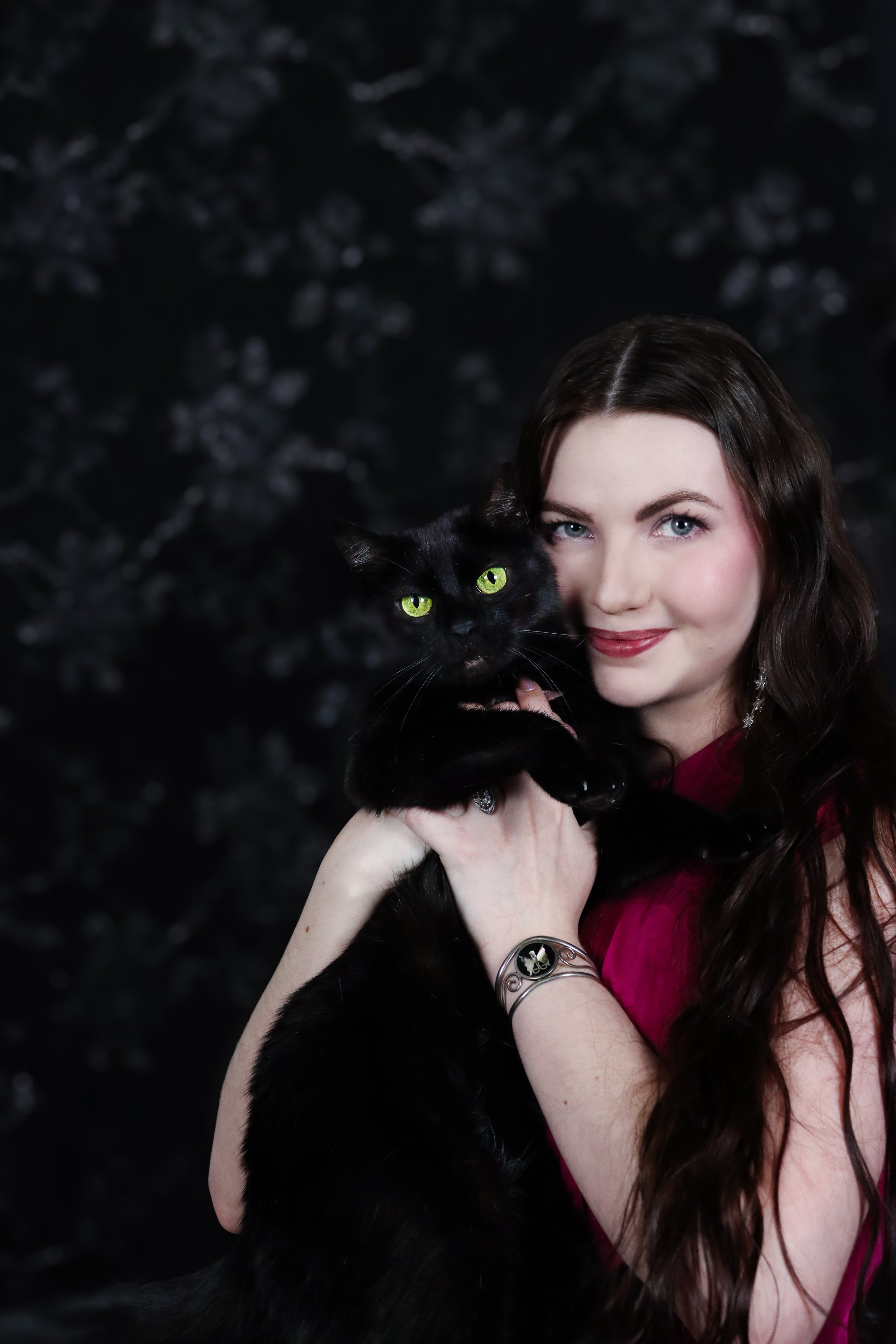 A woman with long dark hair and fair skin holding a black cat with green eyes against a blurred dark background.