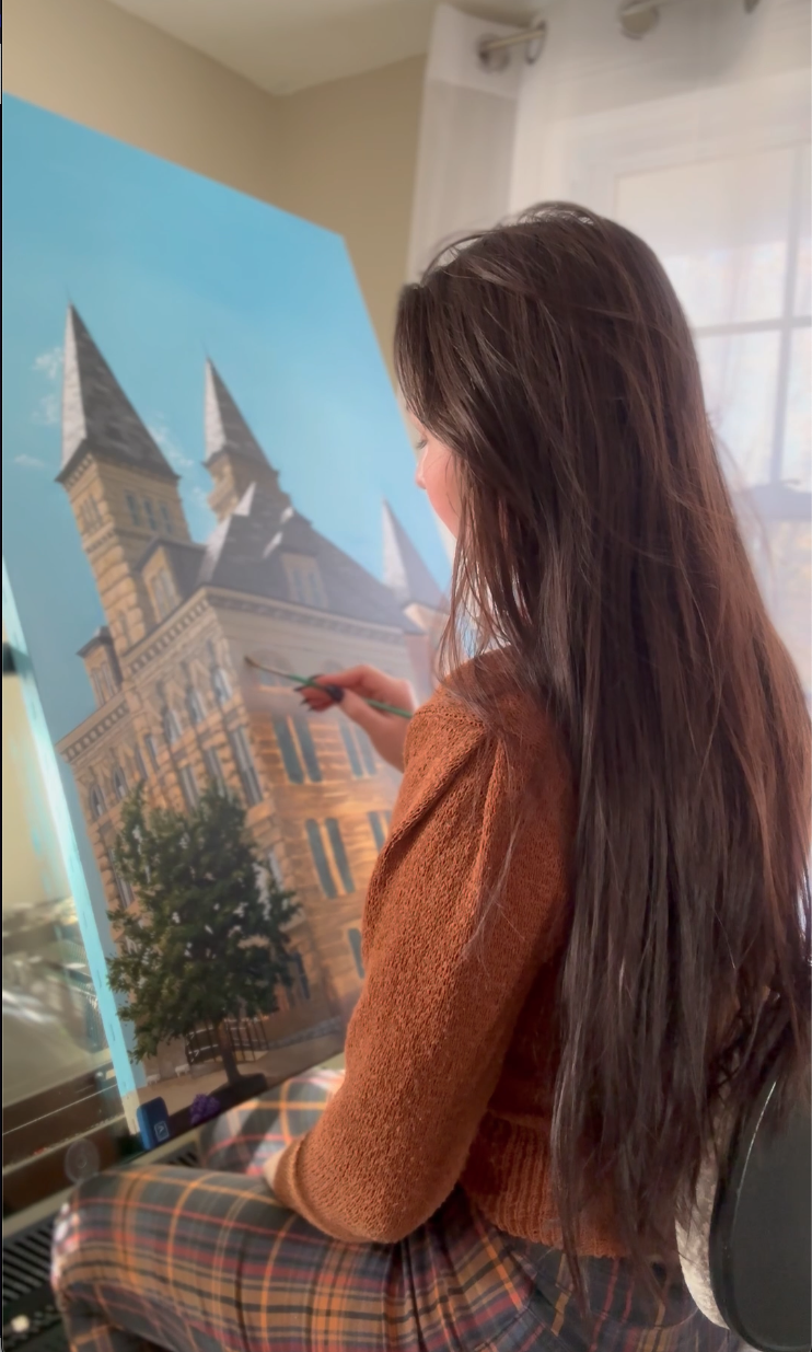 Woman painting a detailed mural of a buliding, trees, and a blue sky.