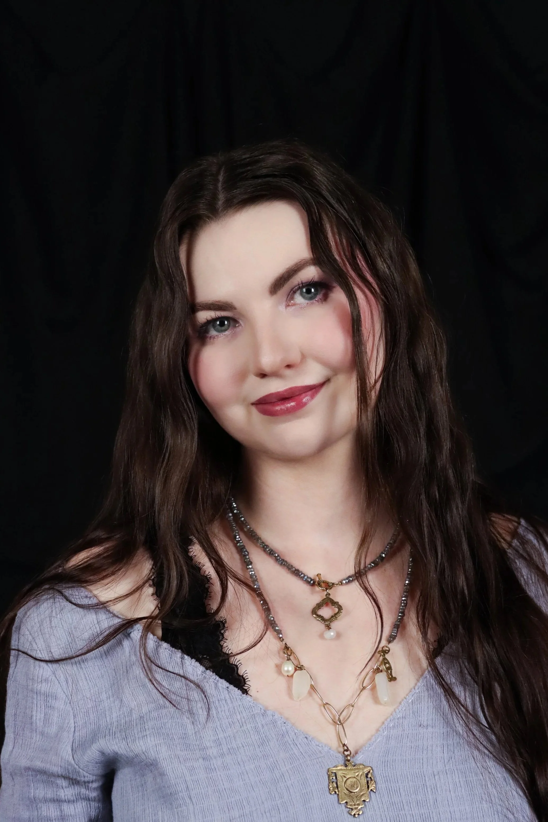 A woman with long wavy brown hair wearing multiple layered necklaces, a Purple top, and a black lace camisole underneath, standing in front of a black background.