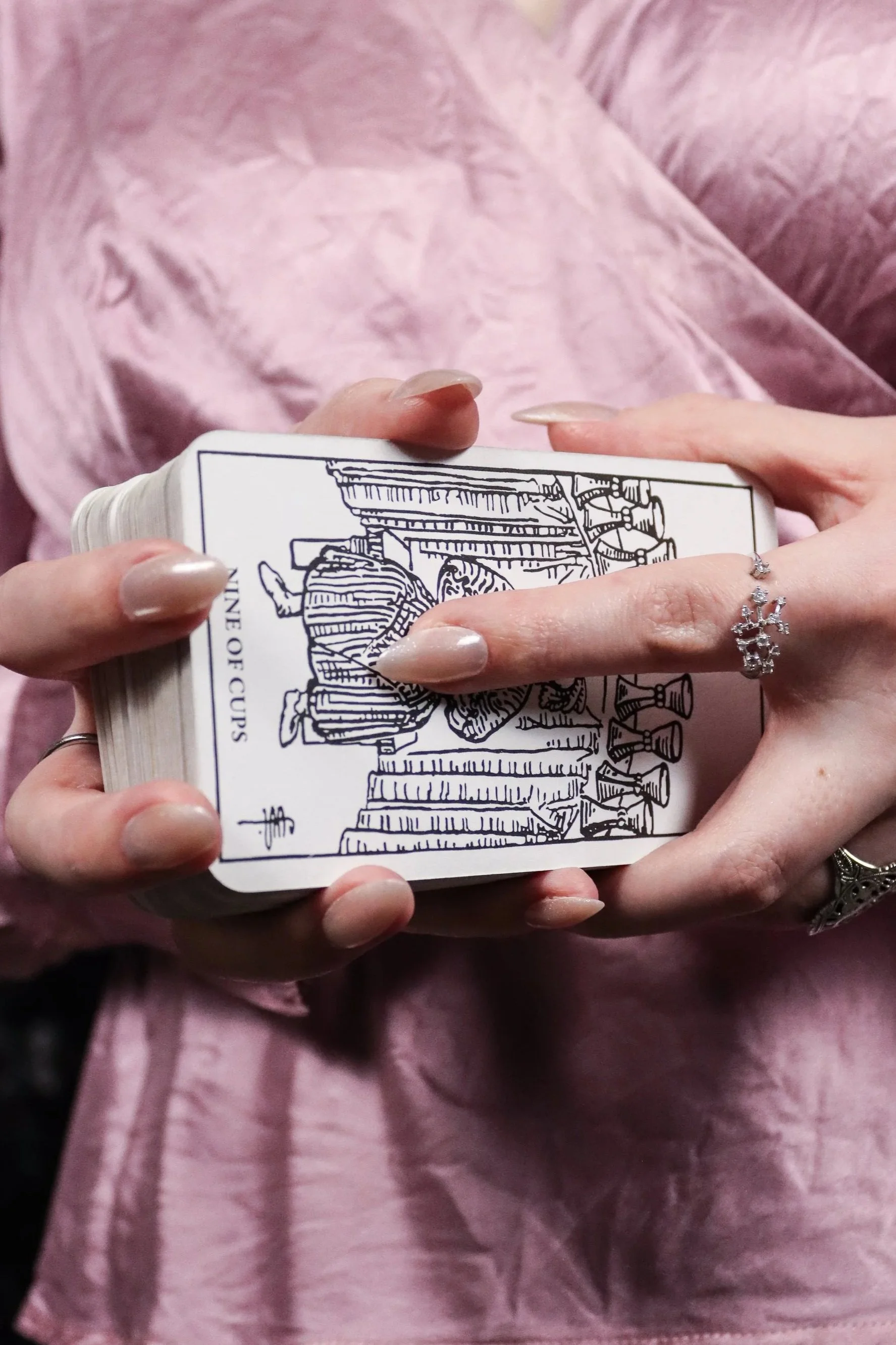 A person with manicured nails holding a deck of tarot cards, displaying the 'Nine of Cups' card, with pink clothing visible in the background.
