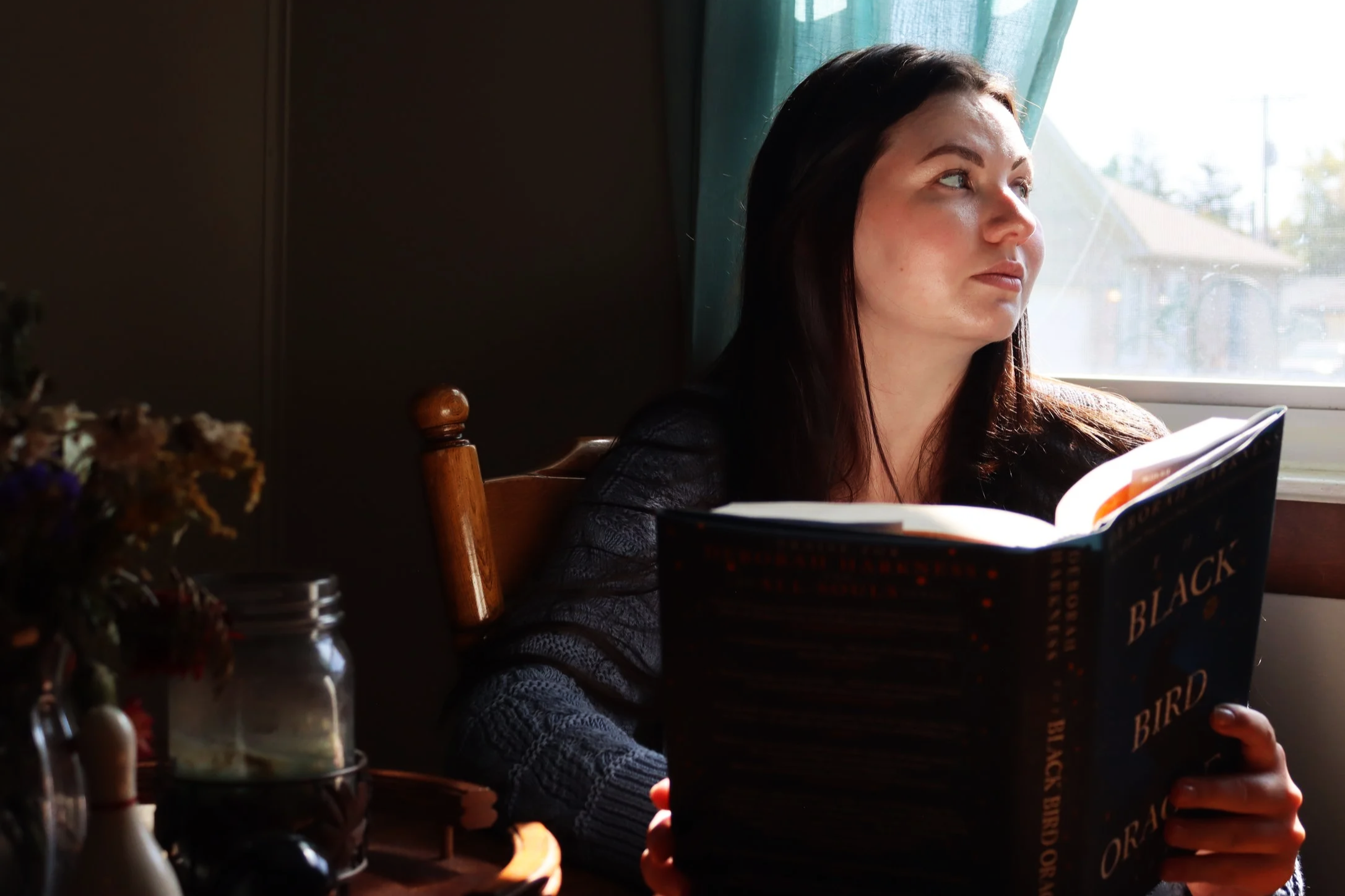 Woman with dark hair reading a book titled "The Black Bird Oracle" near a window.