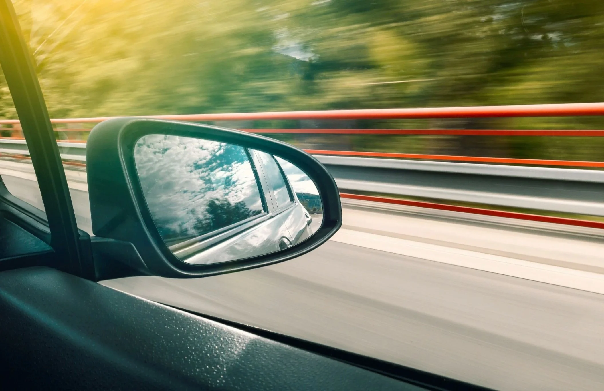 View of a car's side mirror reflecting the sky with clouds while the car is in motion on a highway with green trees and orange safety barriers outside.