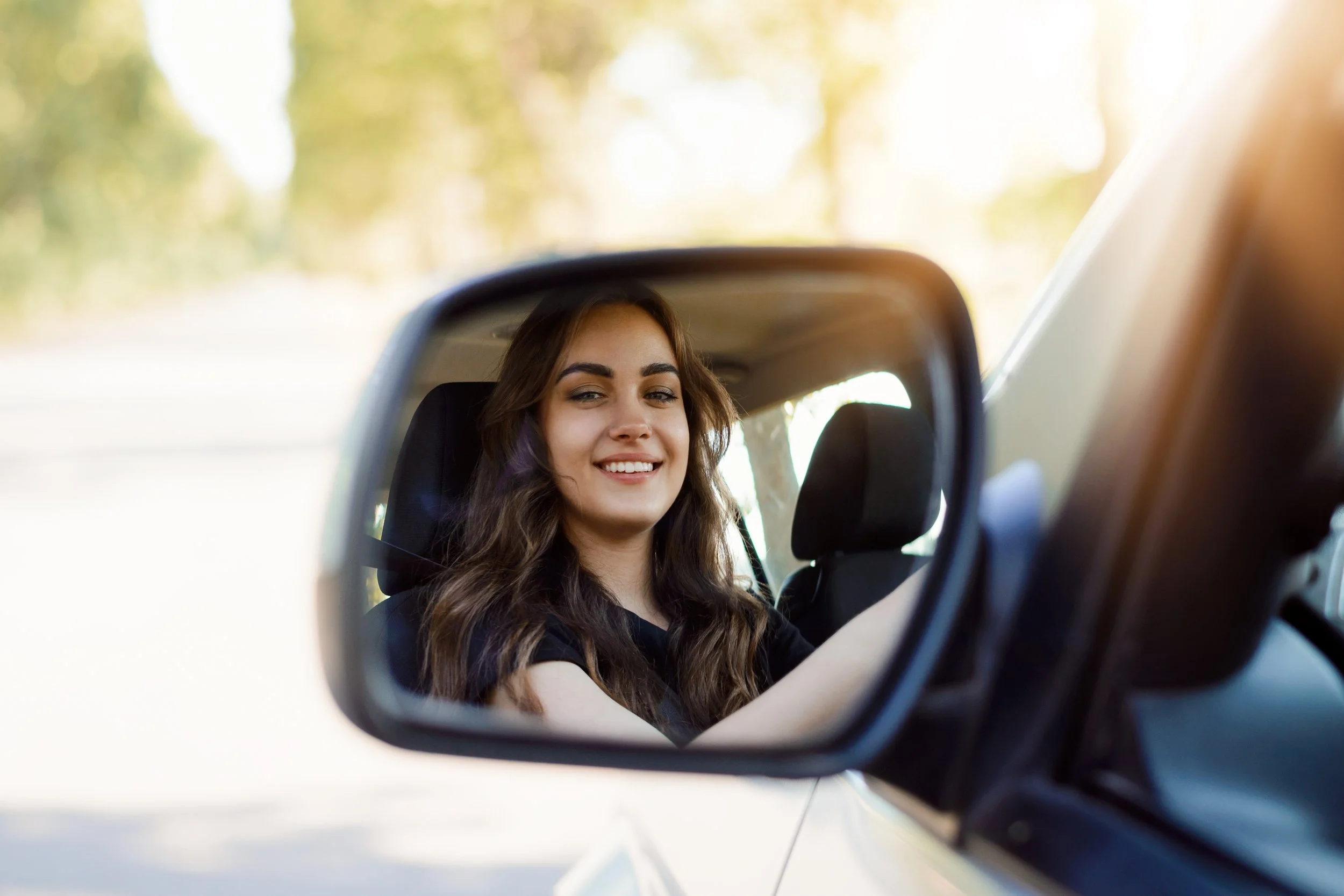 A young woman with long wavy hair smiling while looking into a car's side mirror, with a blurred outdoor background of trees.
