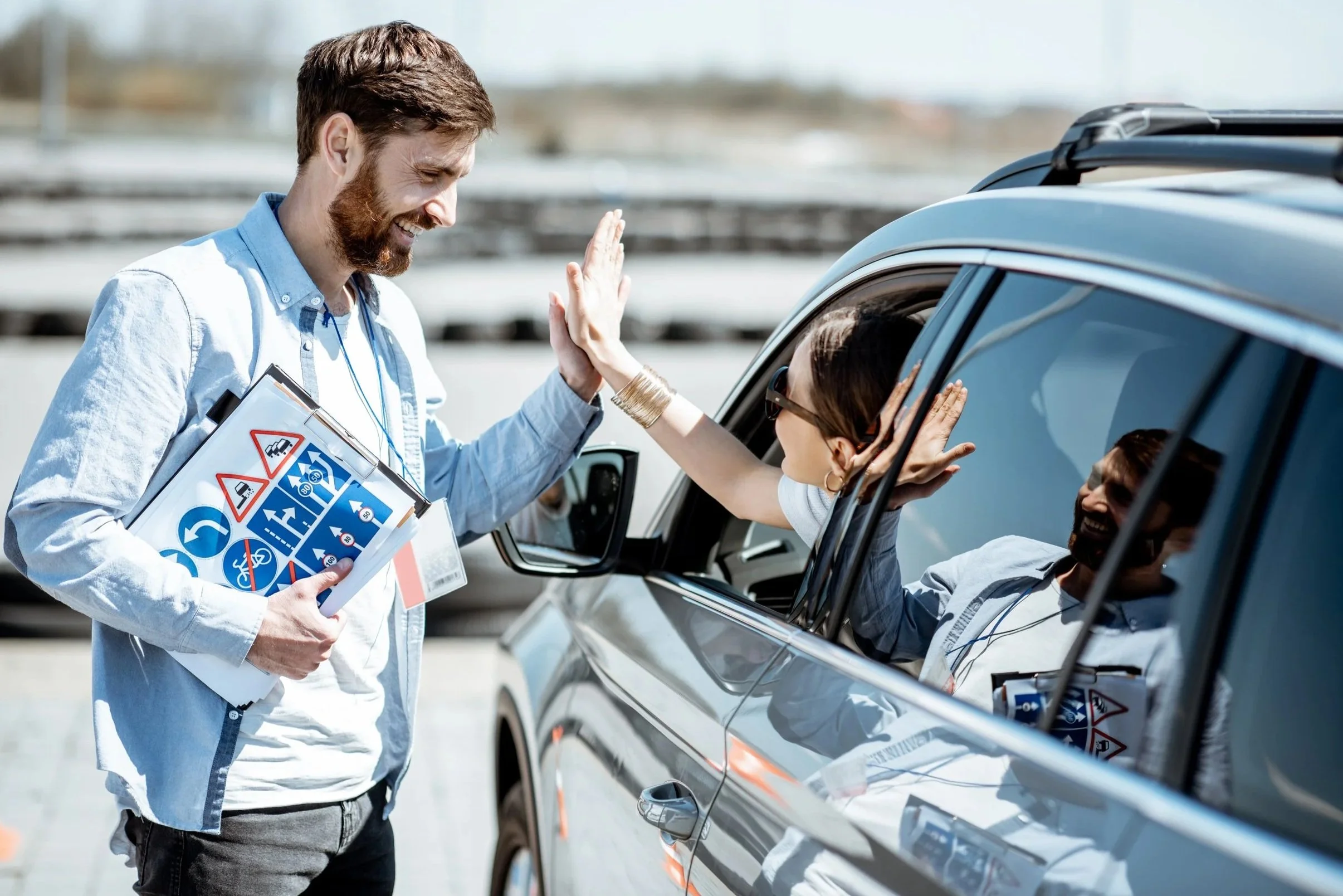 A man and a woman giving a high five through a car window, with a man holding a traffic safety booklet, outdoors