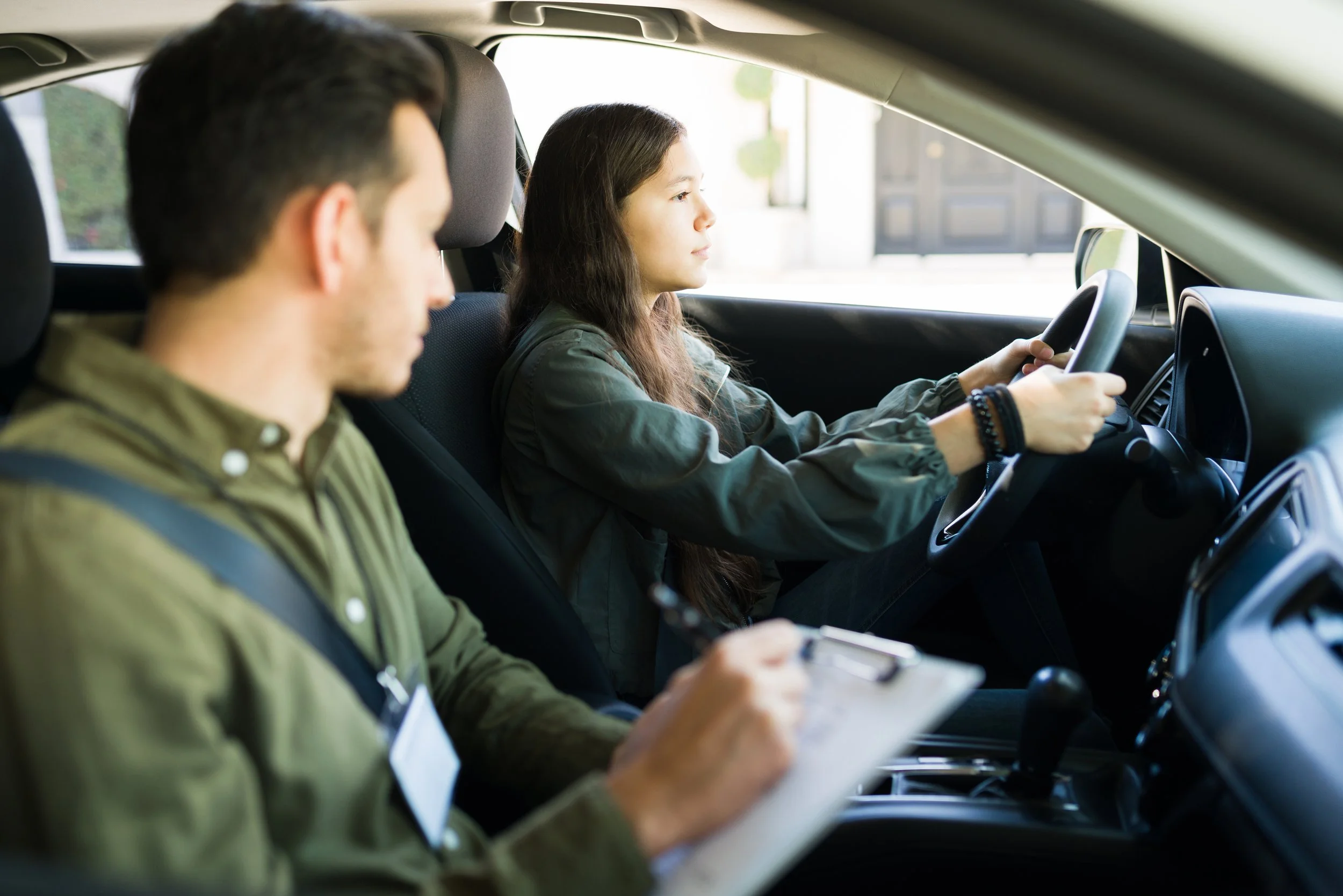 A young woman drives a car while a young man takes notes on a clipboard in the passenger seat.