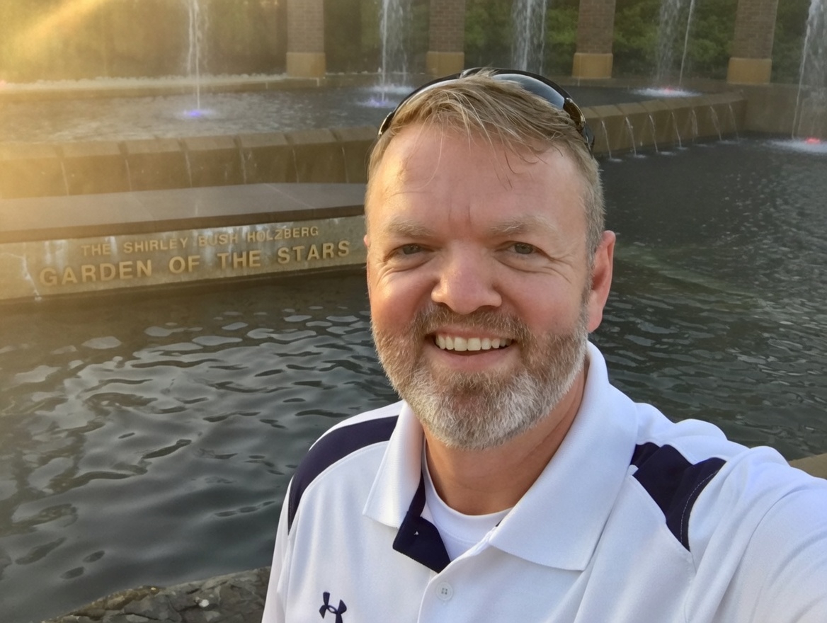Selfie of a smiling man with a beard, wearing sunglasses on his head, in front of the fountain at the Garden of the Stars at the Shirley Bush Holtzberg