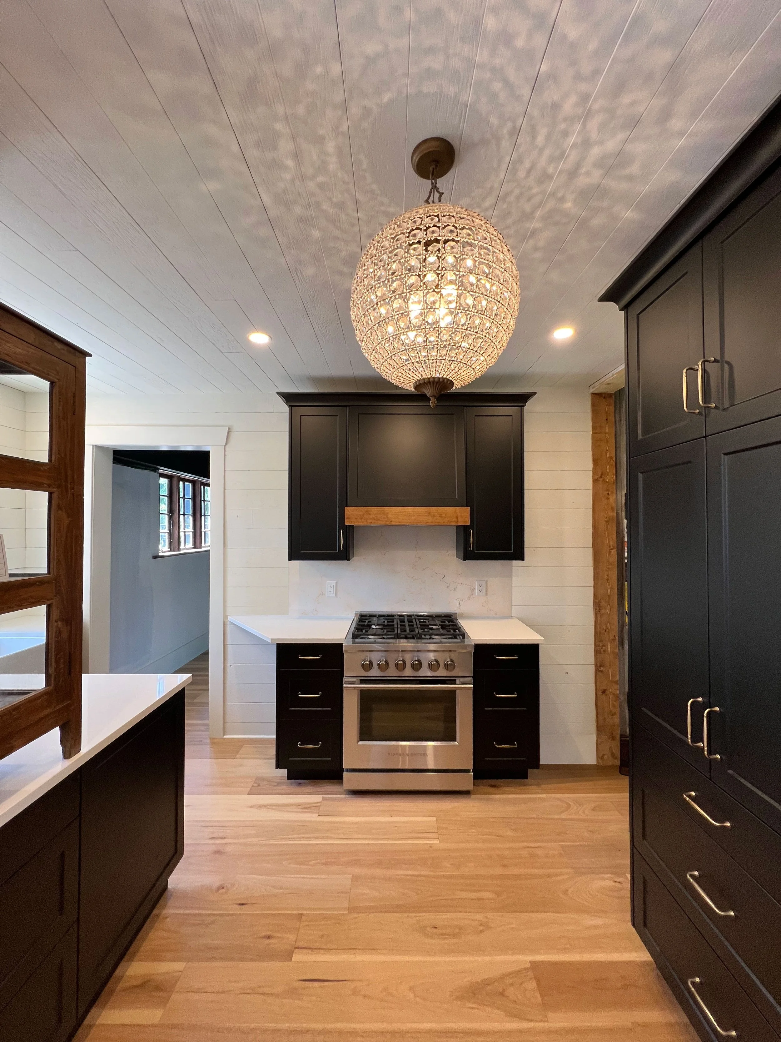 Modern kitchen renovation with black cabinets, a stainless steel stove, light wood flooring, a white countertop, and a round chandelier hanging from the ceiling.