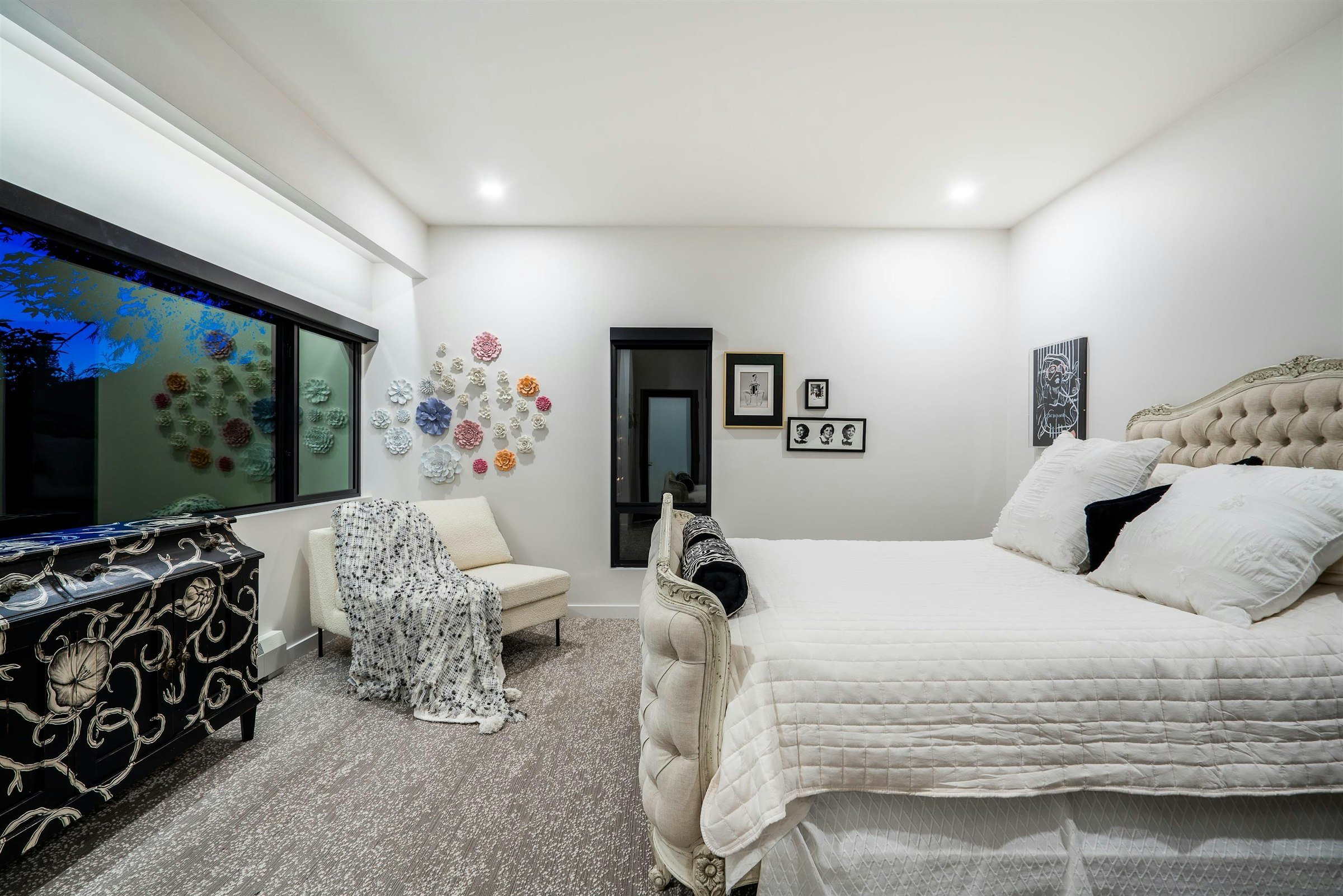 A modern bedroom with white walls and a large bed with an ornate beige headboard. There is artwork on the walls, a window with a dark frame, and decorative items including a black and white dresser and a chair with a throw blanket. The room has a gray carpet.