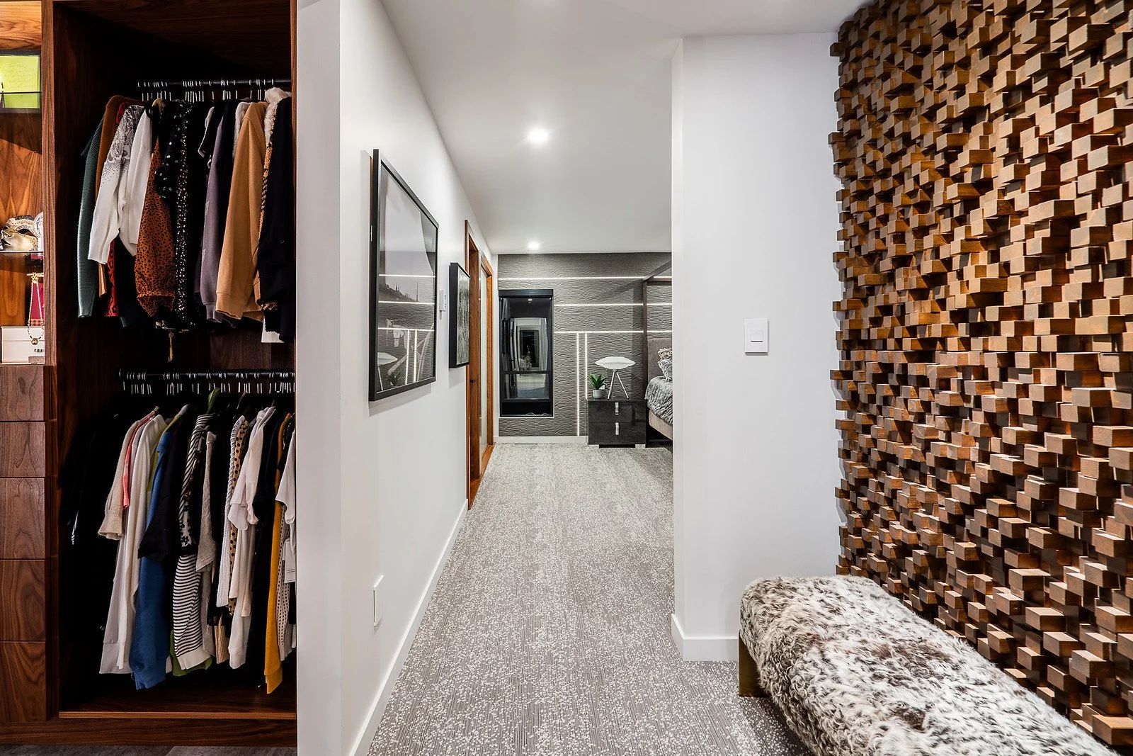 Interior view of a modern home hallway with a walk-in closet on the left, white walls with framed artwork, textured ceiling, and a plaid carpet. At the end of the hallway is a bedroom with a bed, nightstand, and decorative wall panels.