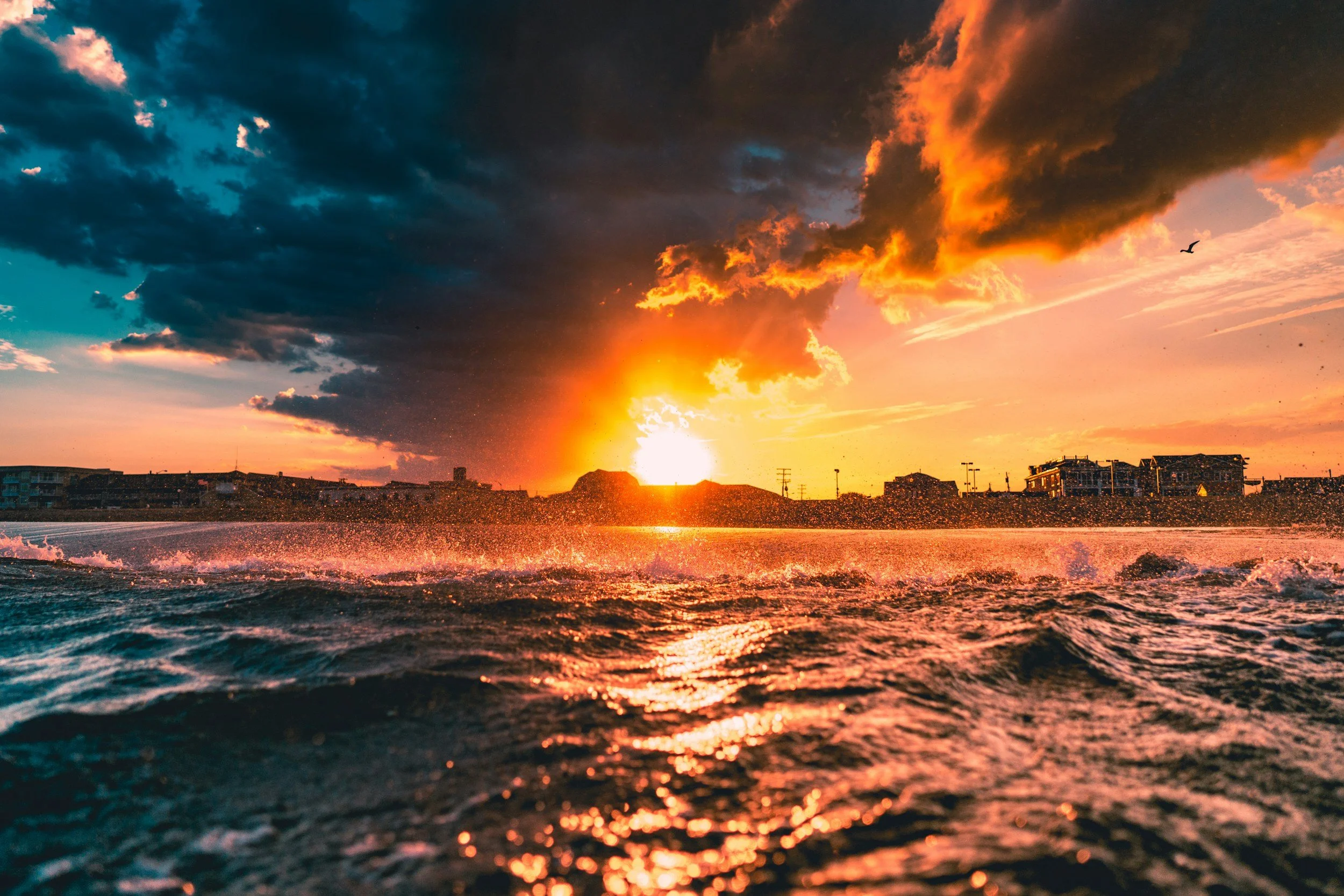 Sunset over the ocean with waves and a city skyline in the background, partly cloudy sky
