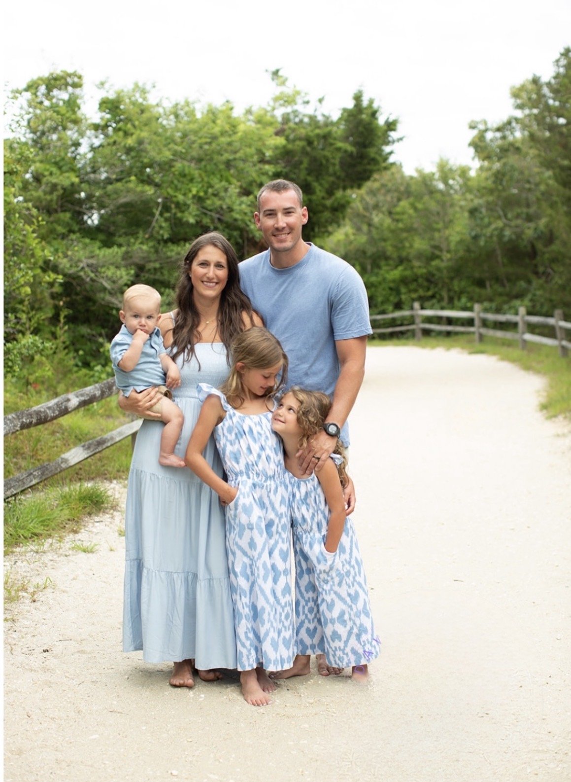 A family of five standing on a dirt path outdoors, surrounded by green trees and bushes. The mother is holding a baby, and two young girls are standing in front of the father, who is smiling and has his arm around one of the girls. All are dressed in light blue and white clothes.