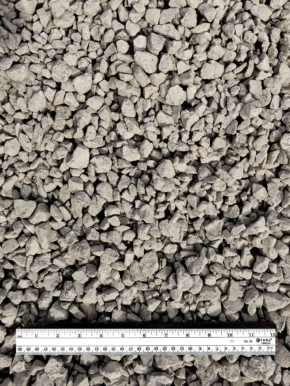A close-up photo of small, irregularly shaped gray gravel stones with a measuring ruler placed across the bottom of the image.
