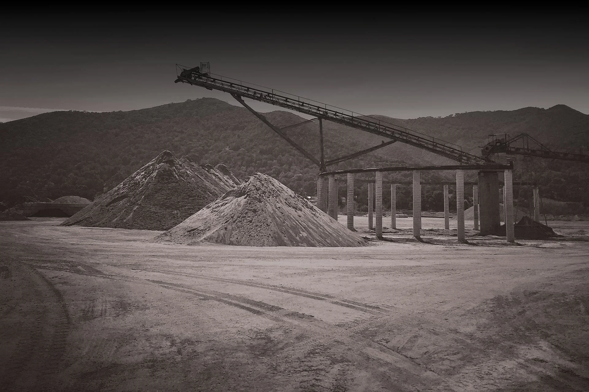 A black and white photo of a salt plant with piles of salt and conveyor belts on support columns against a mountainous background.