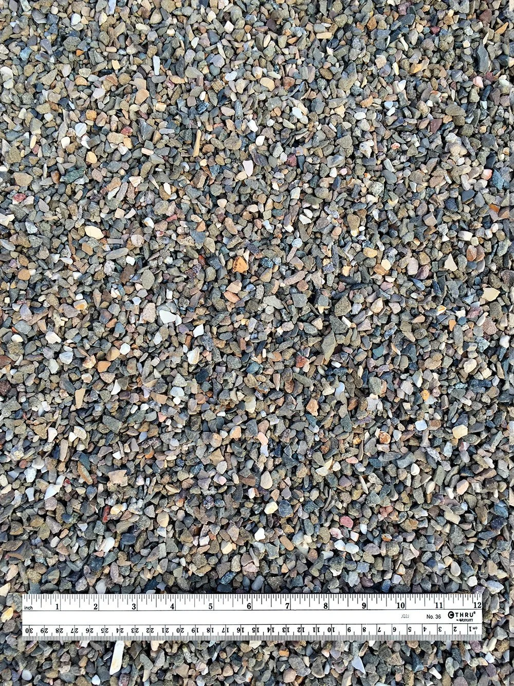 Close-up view of a gravel surface with a measuring ruler at the bottom, showing gravel stones of various small sizes and colors.