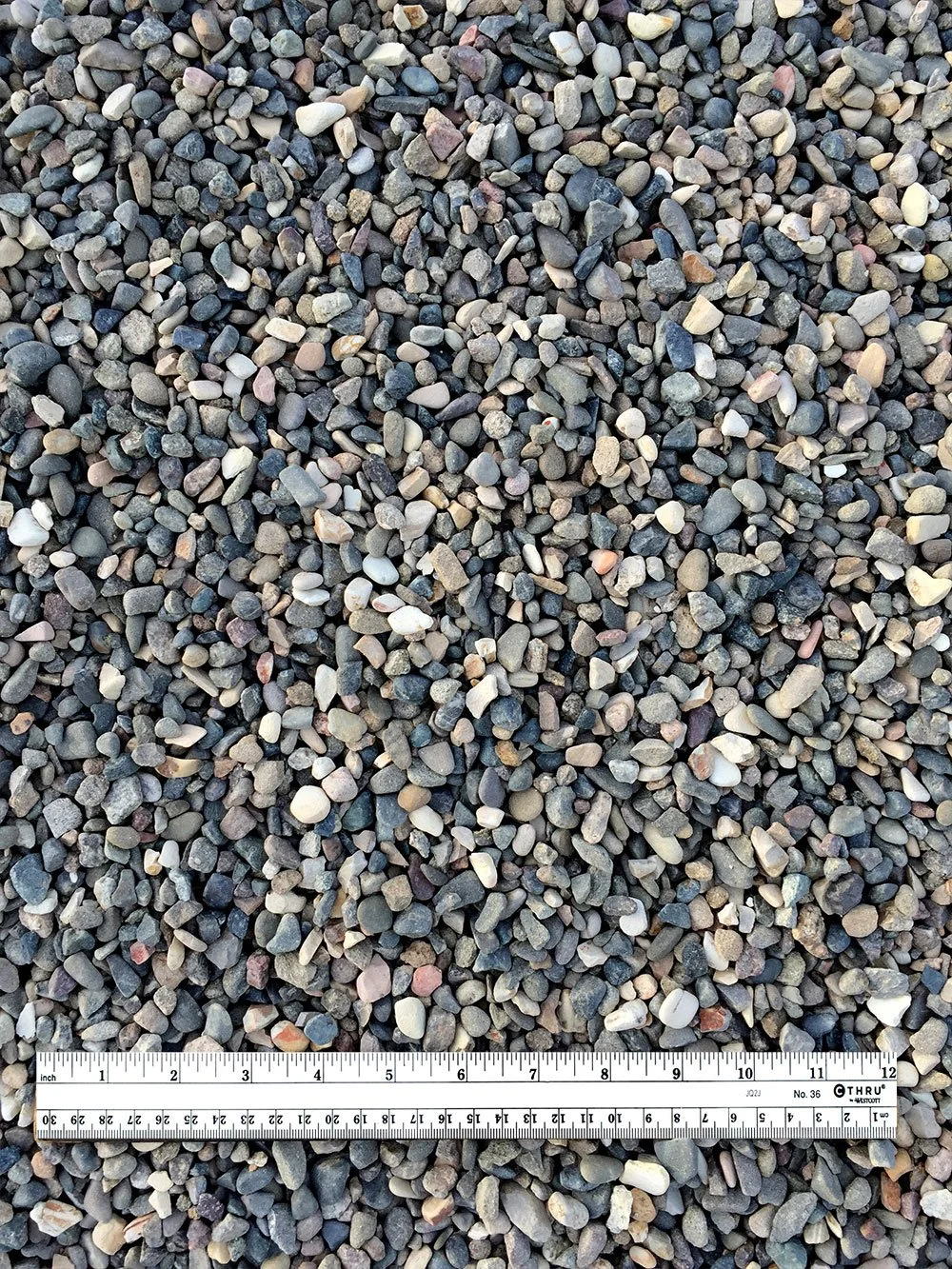 Close-up view of small multicolored gravel stones with a measuring ruler placed across the middle, showing centimeters and inches.