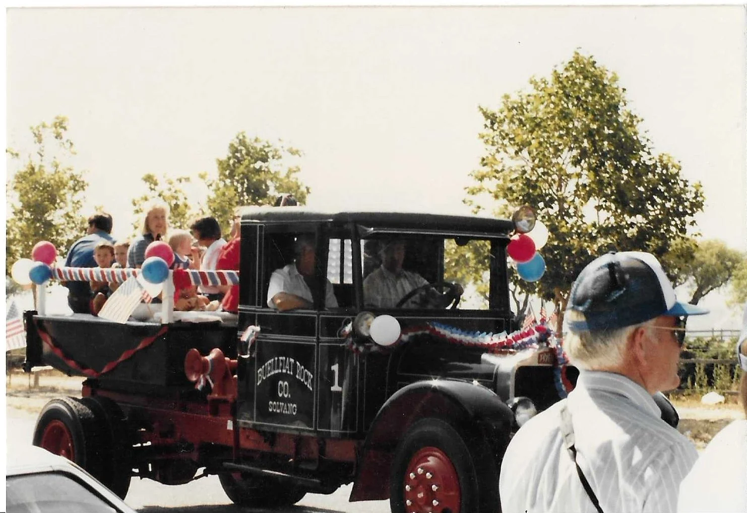 Vintage truck decorated with red, white, and blue bunting and balloons, carrying parade spectators during a patriotic celebration, with trees and a clear sky in the background.