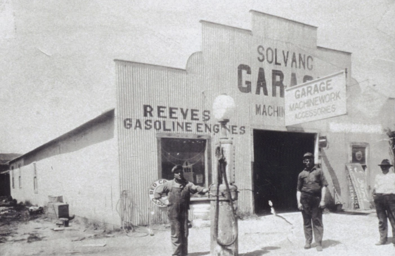 Black and white photo of a vintage garage with three men standing outside. The building has signs for gas, machinery, and accessories, with a large sign reading 'Solvang Garage'. One man is leaning against an old-fashioned streetlamp, and another is holding a hanging animal carcass.