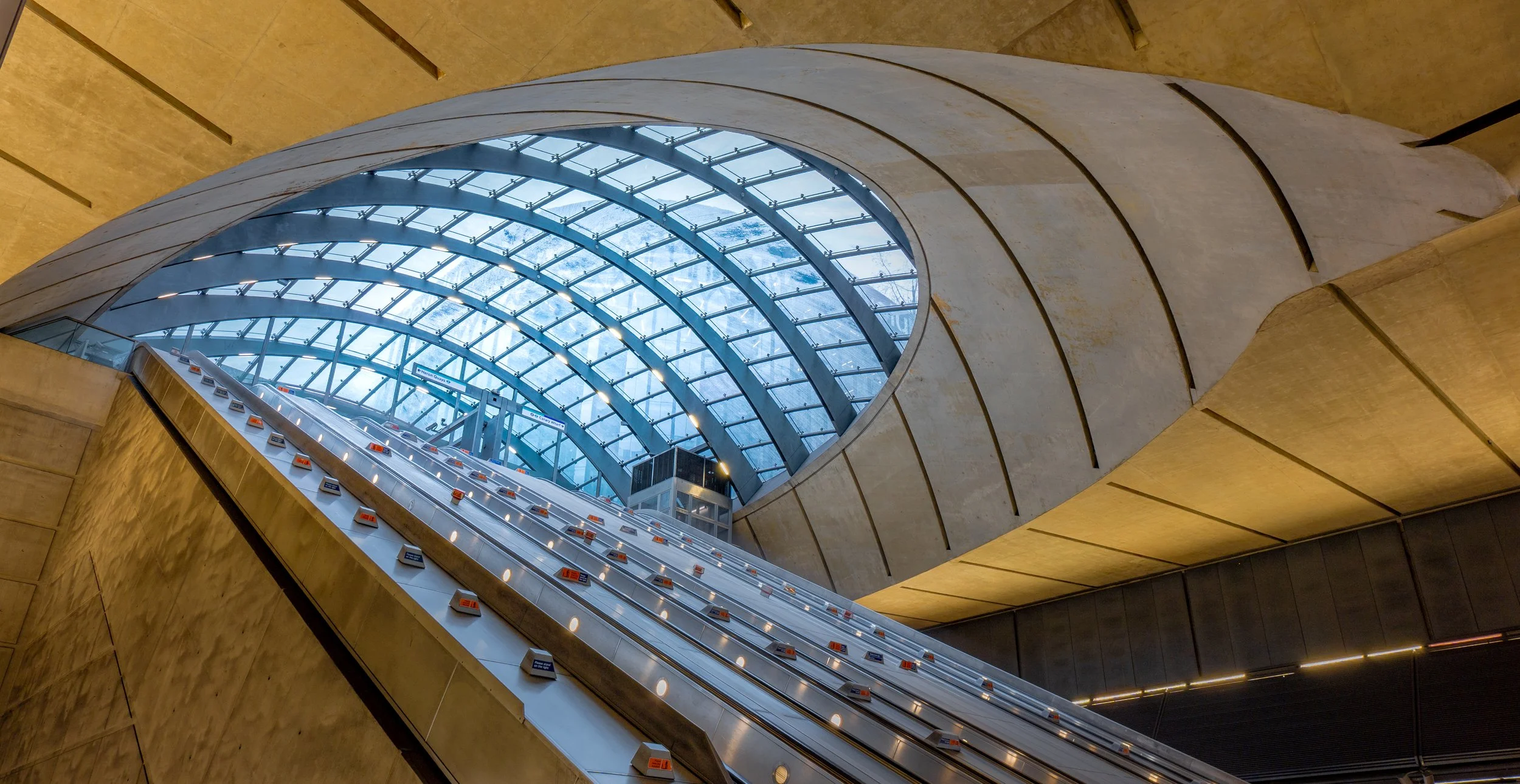 Interior of a modern building with a large, curved glass ceiling and an escalator leading upward, surrounded by gold-colored walls.